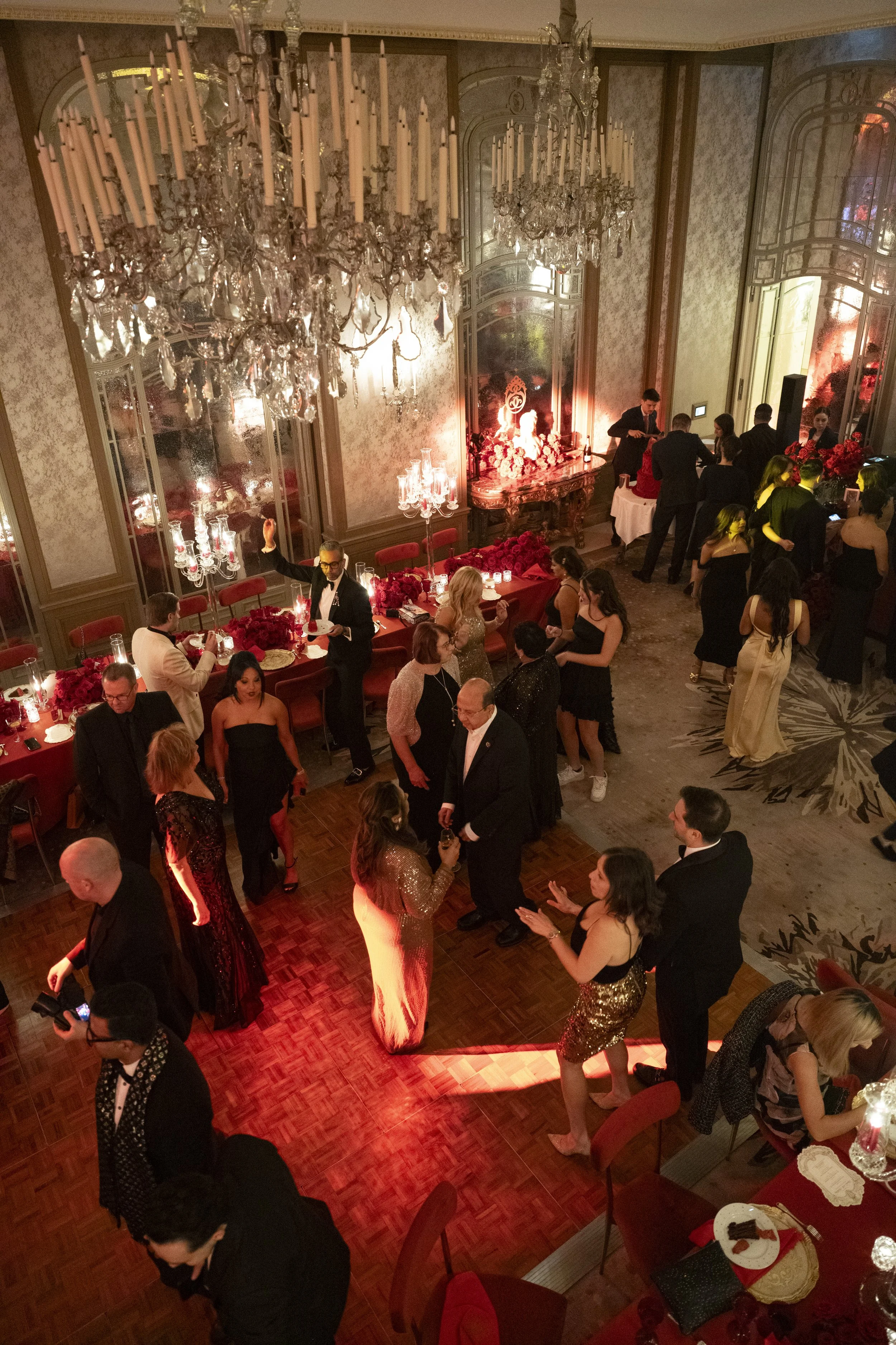 A festive indoor party scene with guests dressed in formal attire, socializing, and dancing in a grand room with chandeliers, ornate mirrors, and floral decorations.