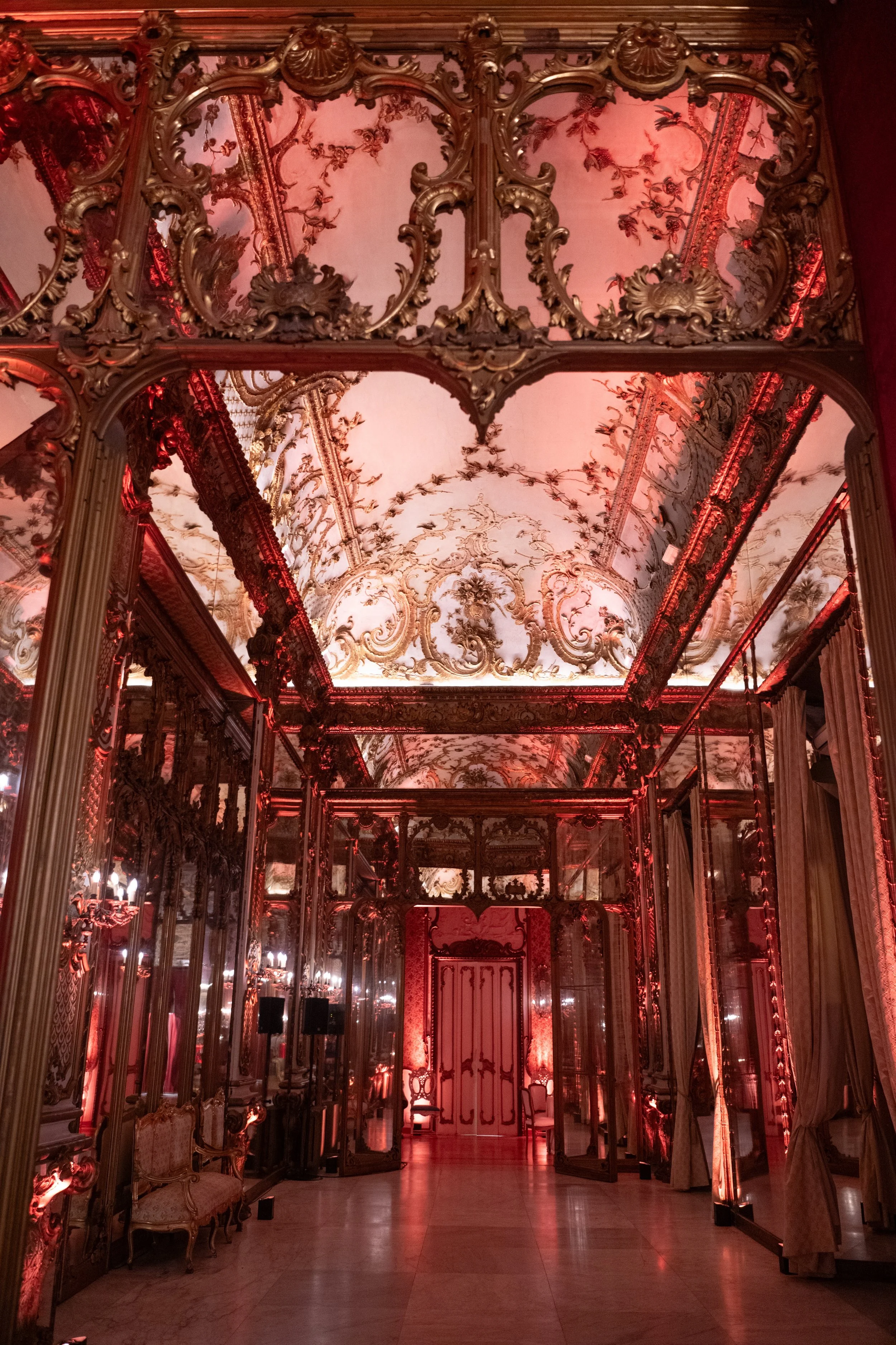 An ornate room with elaborate gold and pink baroque-style decor, featuring mirrors, mirrors, and intricate moldings, illuminated with warm red lighting.