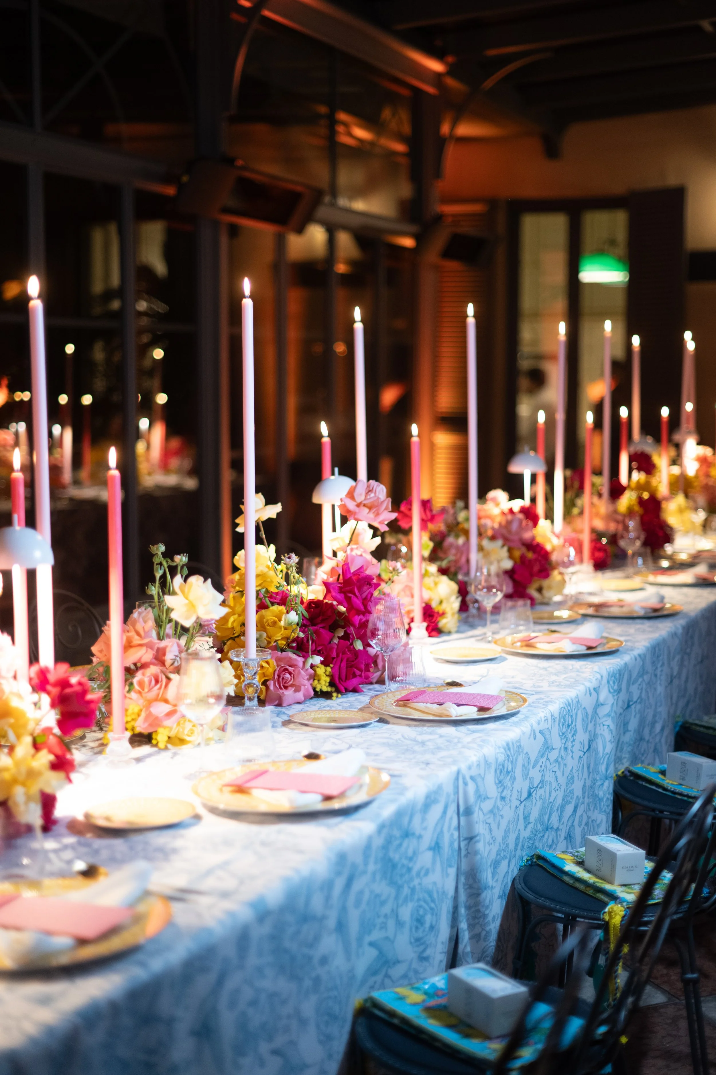 A long banquet table set for a celebration with many pink and yellow flowers, tall pink and white candles, place settings with plates, napkins, and glasses, in an elegant indoor venue with dark wood accents and warm lighting.