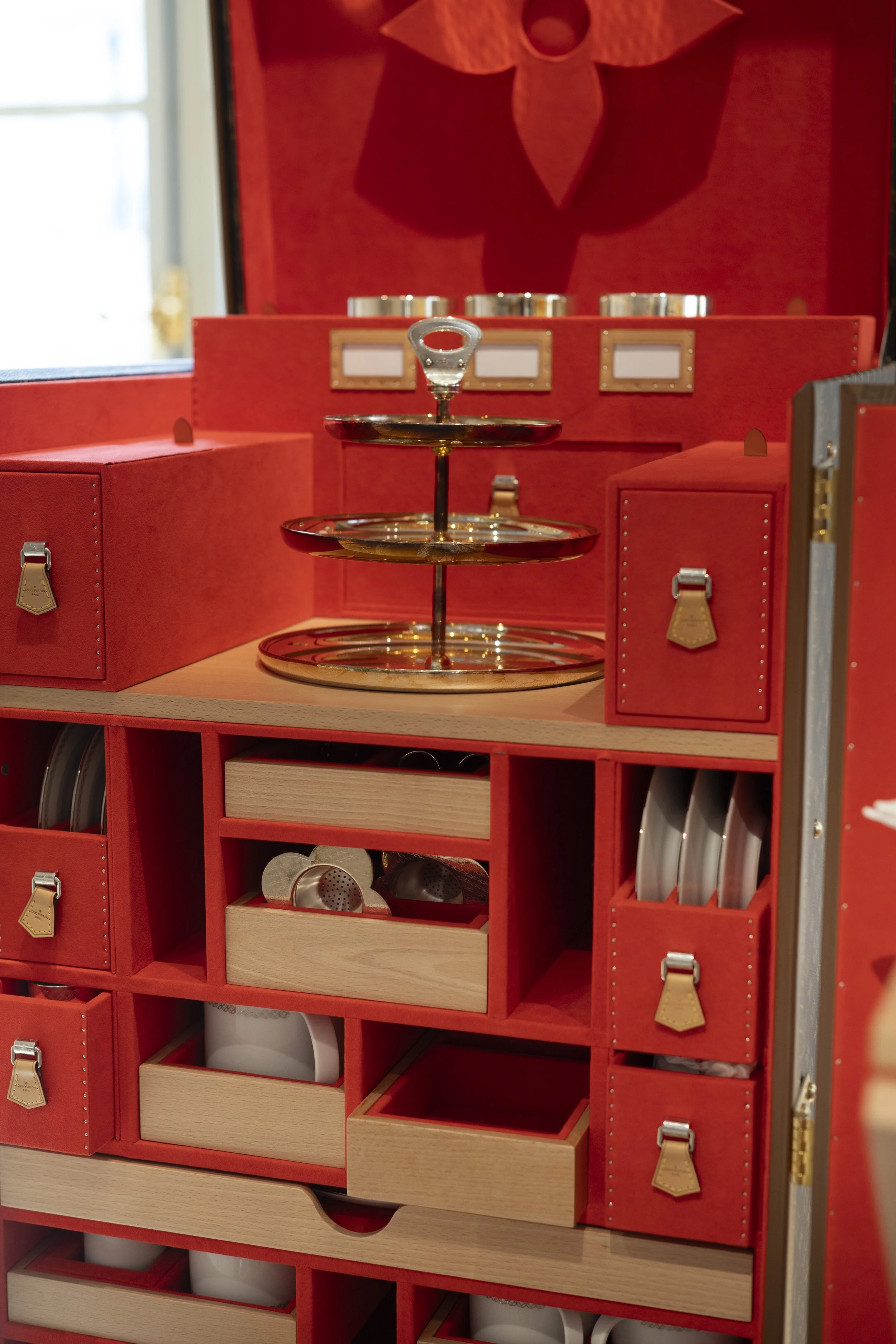 Red cabinet with various compartments containing kitchen utensils, including a tiered serving tray, metal containers, a white mug, and kitchen tools.