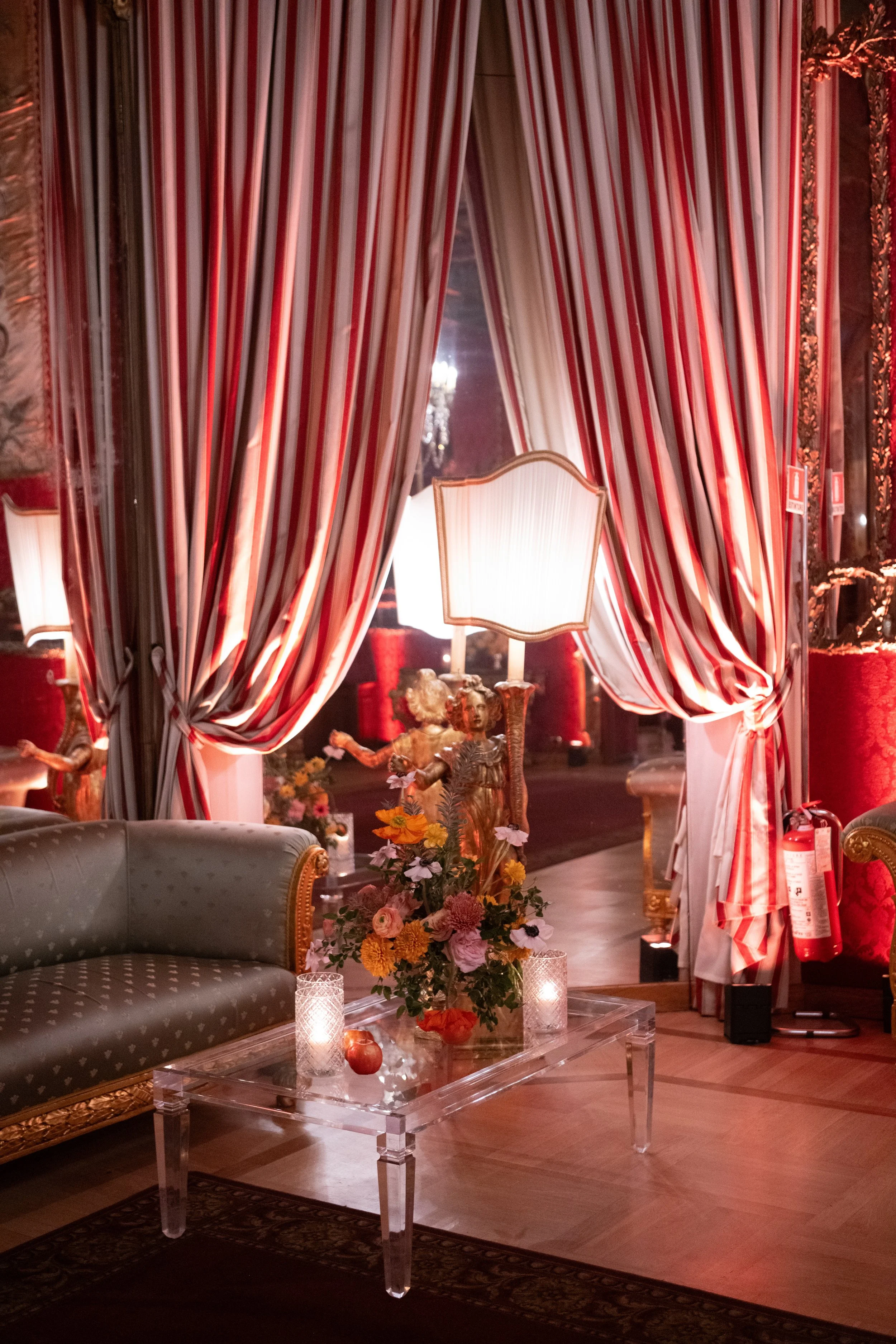 Elegant room with draped red and white striped curtains, a floral centerpiece on a transparent table, a beige sofa with gold accents, candles, and a chandelier reflected in the mirror.