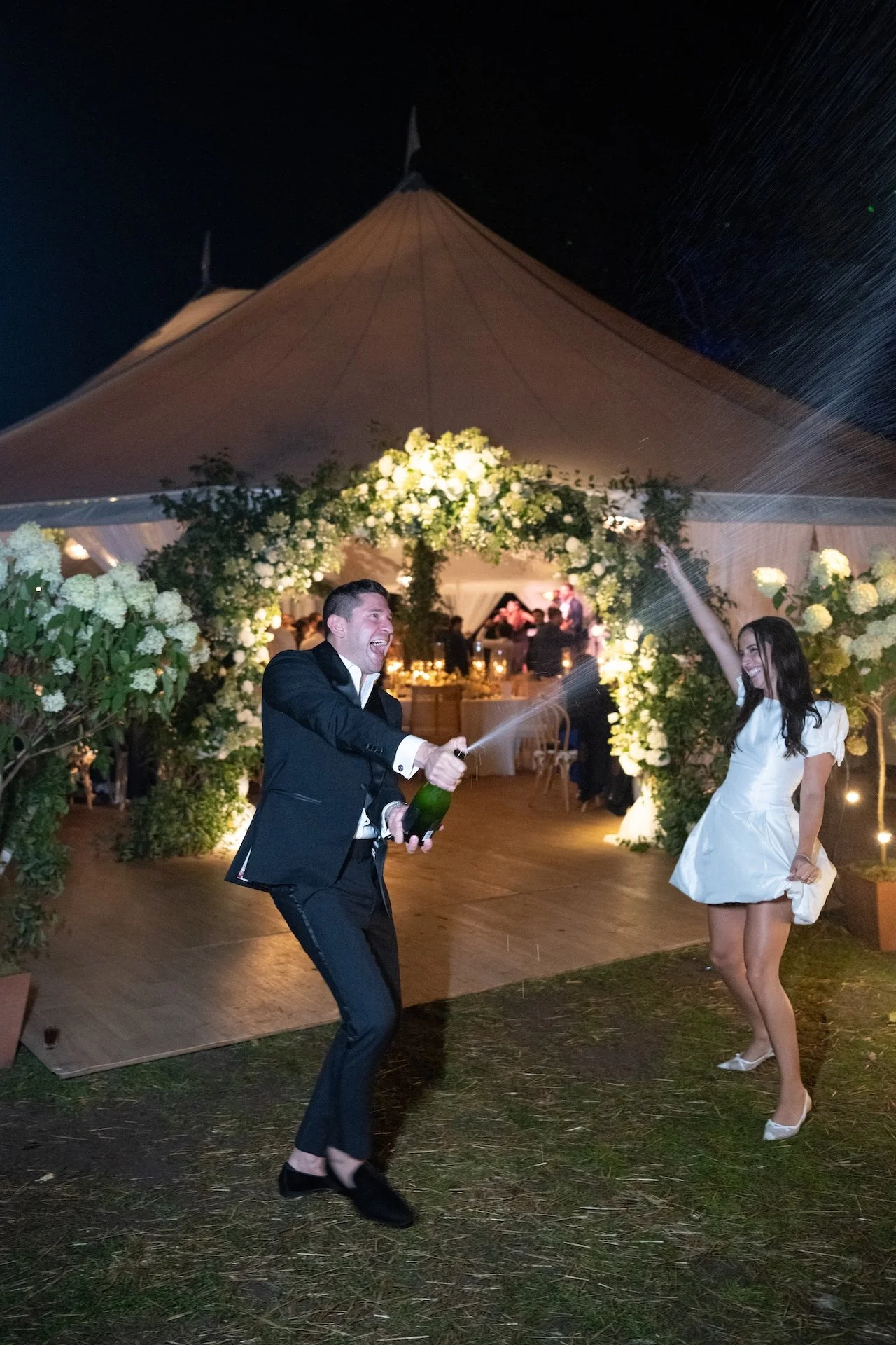 A man and woman celebrating at a wedding reception, with the man spraying champagne and both smiling, under a large decorated tent at night.