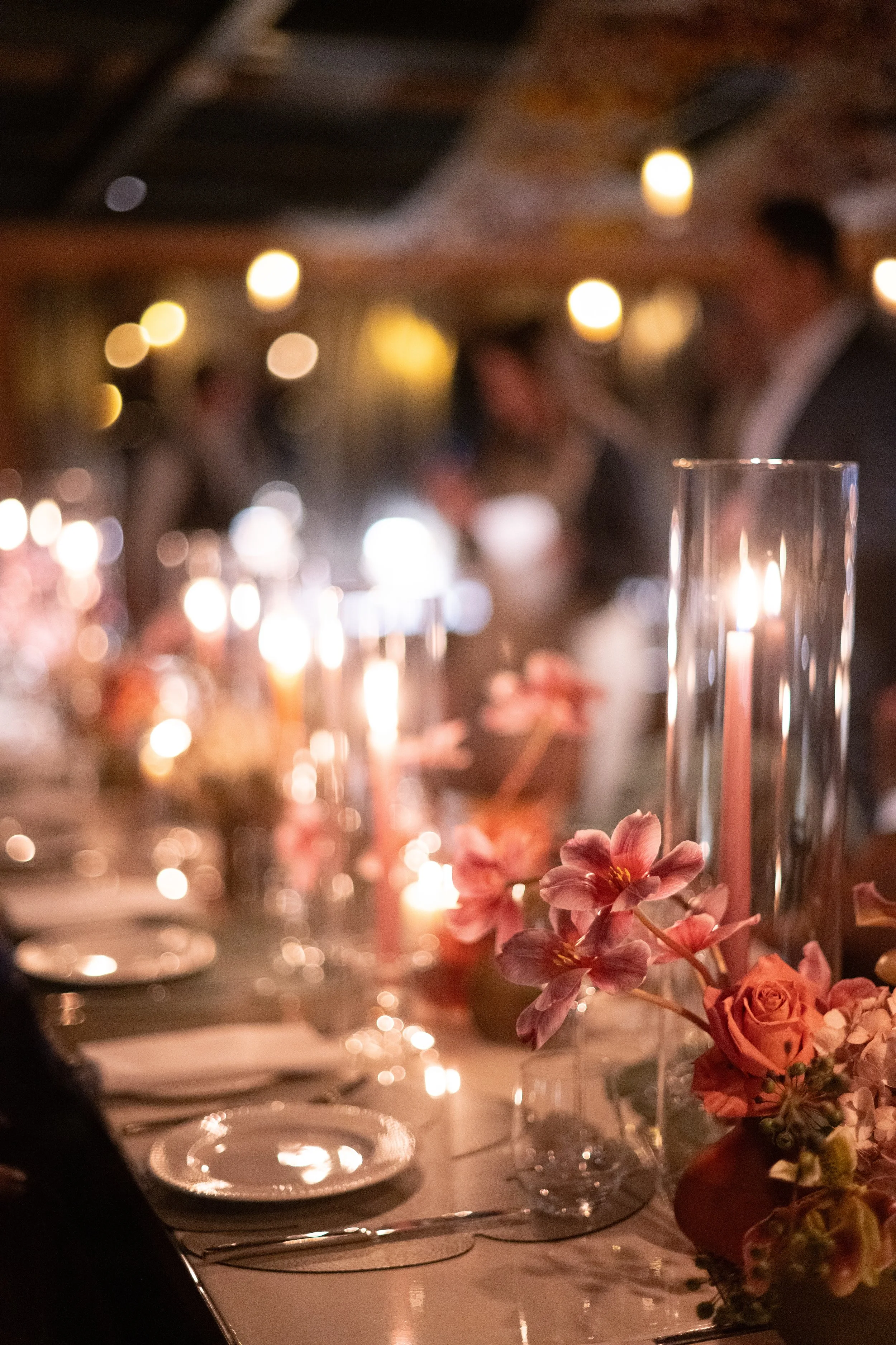 A table decorated with pink flowers, candles, and elegant tableware, set in a dimly lit room for a formal event or dinner party.