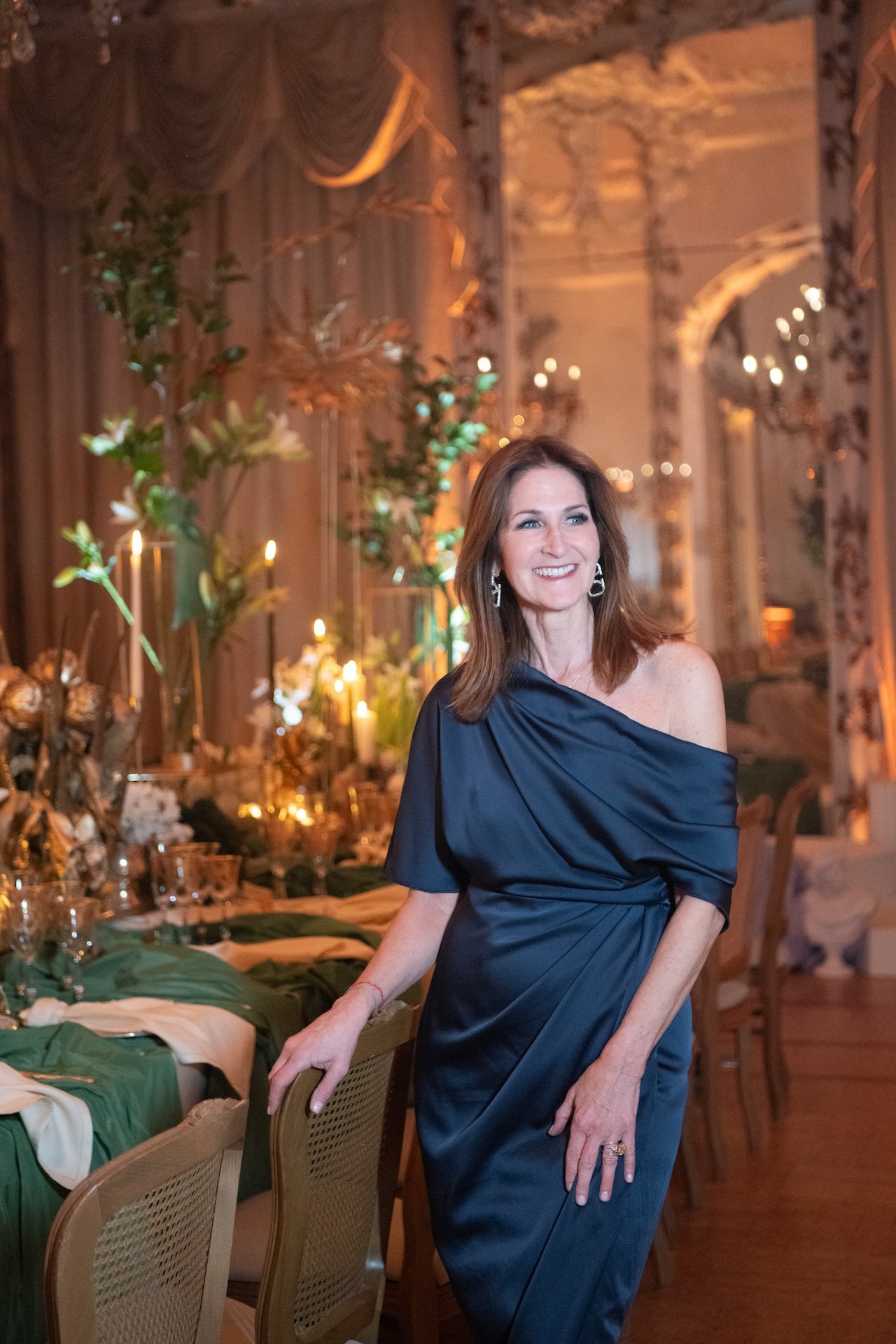 A woman in a navy blue satin off-shoulder dress smiling at an elegant, warmly lit event with decorated tables, greenery, and chandeliers.