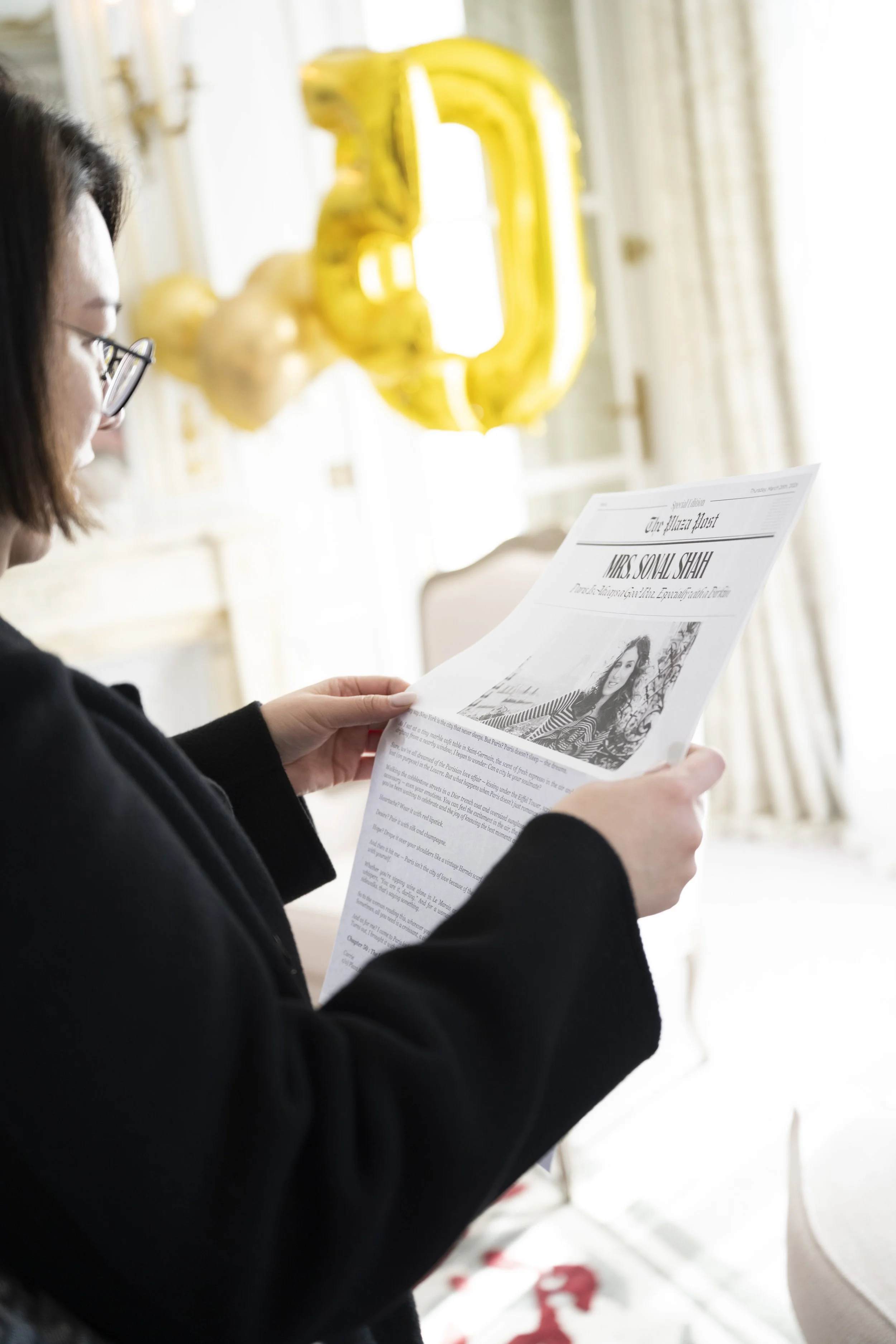 A woman wearing glasses and a black sweater is reading a newspaper with a prominent headline about Miss Sowal Shah, in a bright room with gold foil balloons shaped as the number 3 and a background of cream-colored curtains.