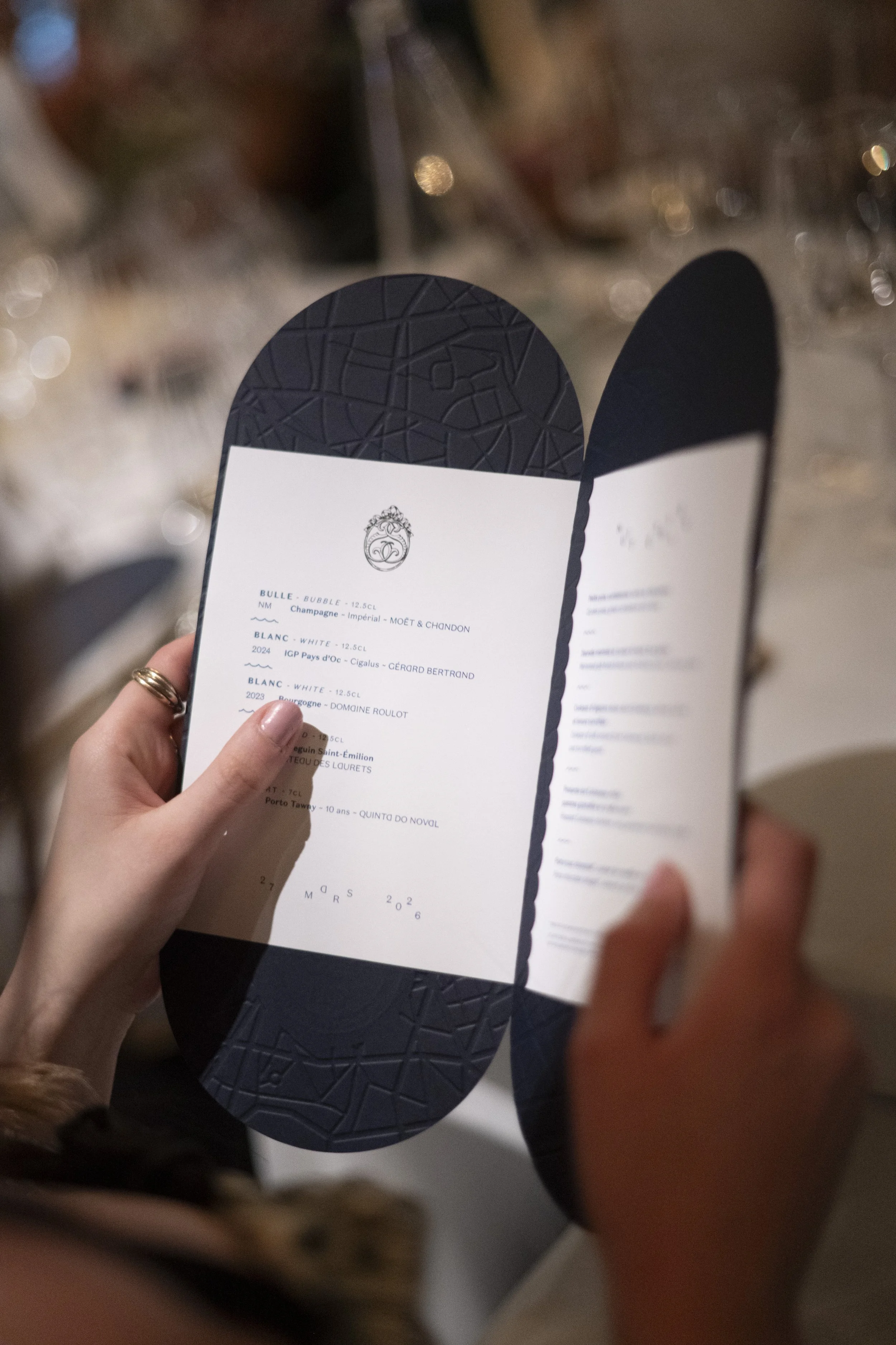 A person holding a black-bordered menu with white paper inside, listing various wines and their details. The person has a ring on their finger and is standing near a blurred background with lights.