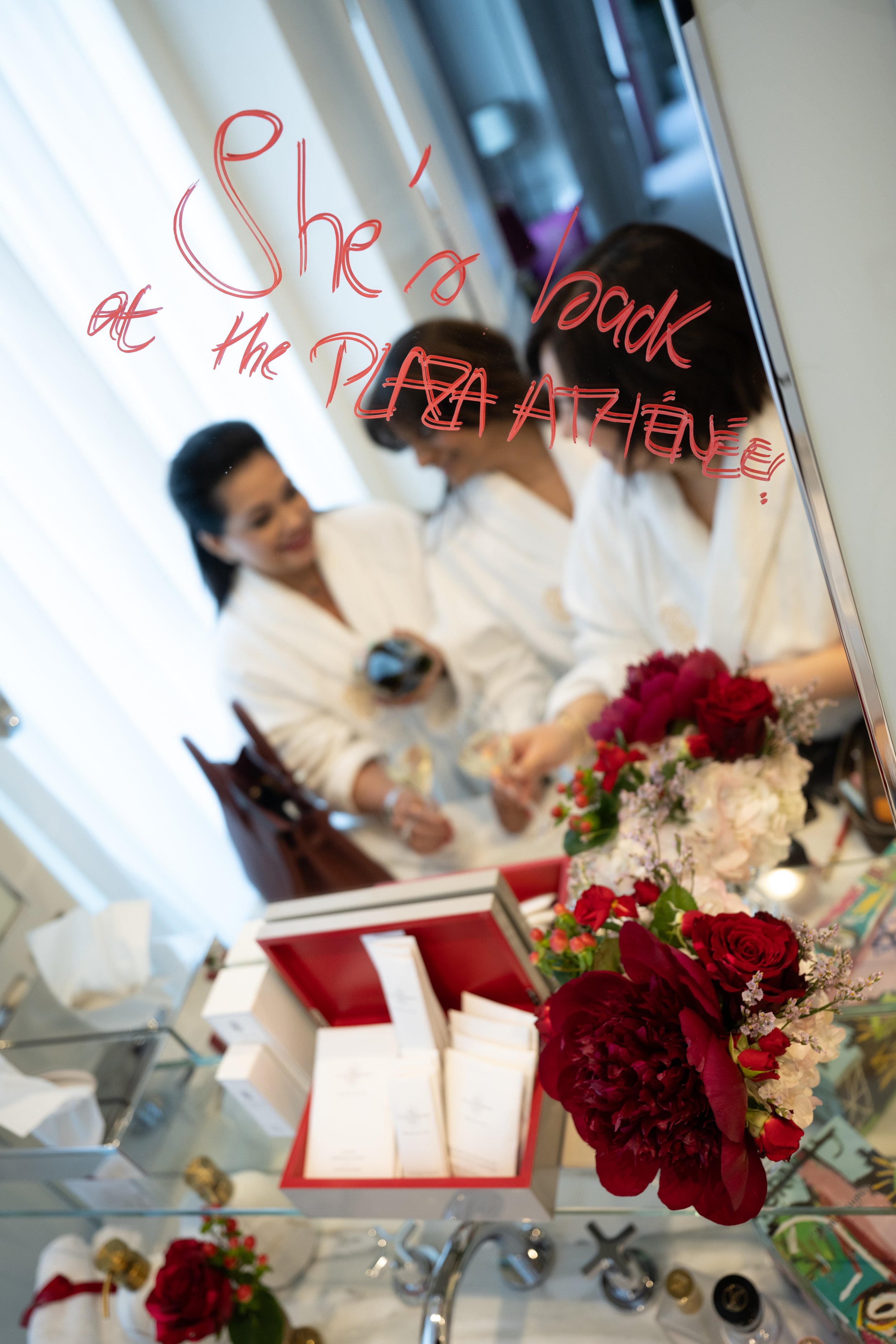 A mirror with red handwritten text that reads 'She is my greatest'}) in a room with women dressed in white robes, and a table with a bouquet of red flowers.