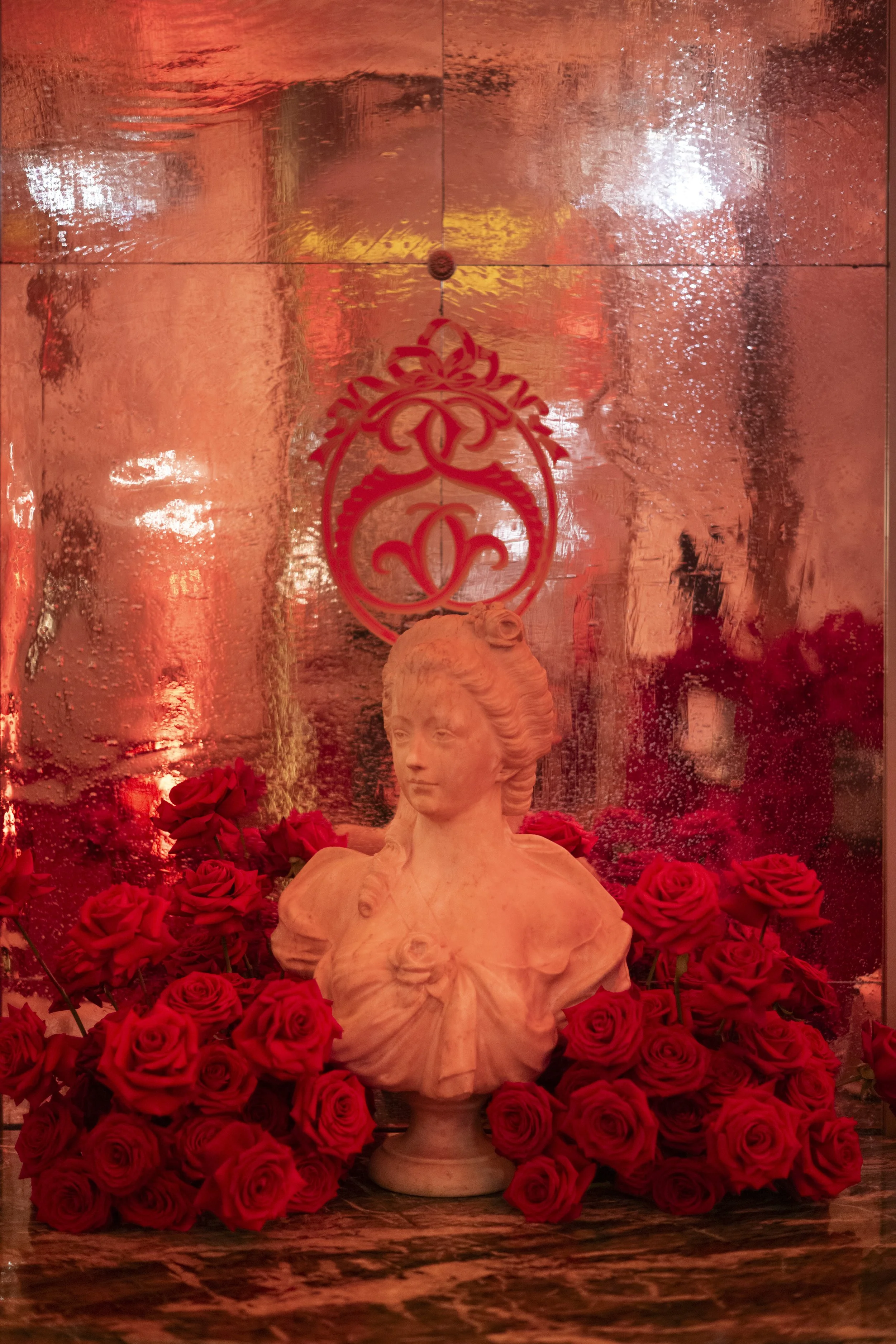 A bust sculpture of a woman surrounded by red roses, with a reflective background and ornate red emblem hanging above.