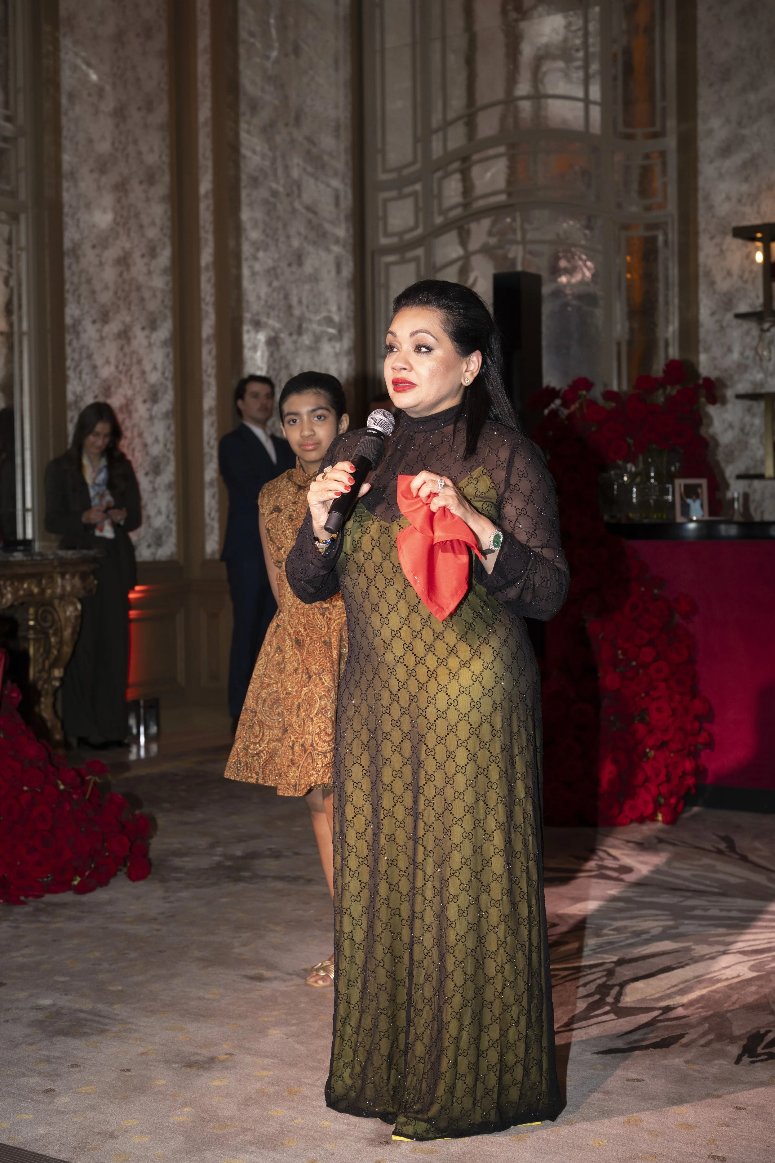 A woman dressed in a long dress with a black lace top and an olive green pattern, holding a red cloth and a microphone. She has dark hair and is standing on a decorated stage with red flowers and a marble wall background. There are three other people