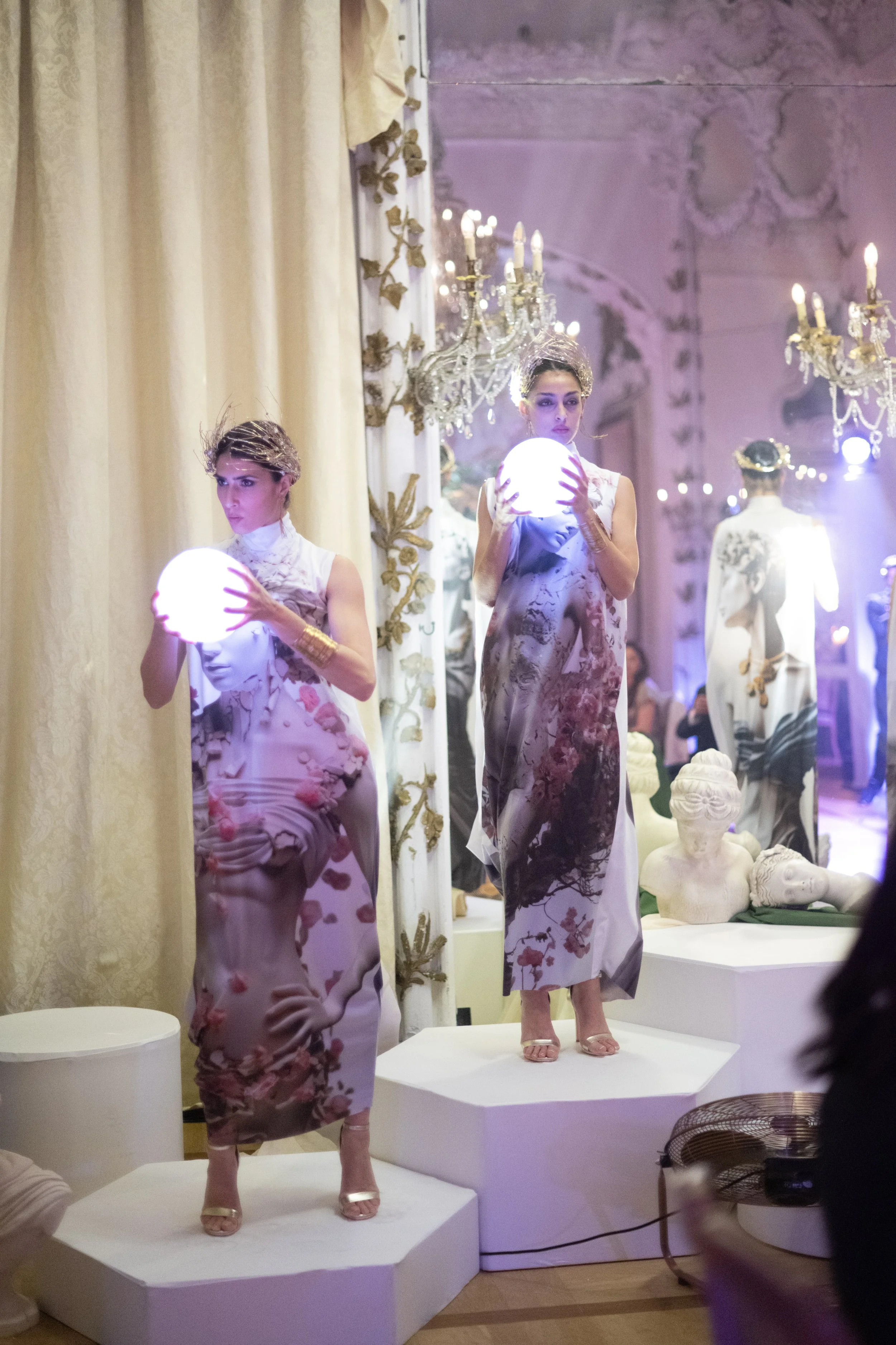 Two models on display stands showcasing floral patterned dresses in an ornate, chandelier-lit room with classical sculptures and decorative elements.