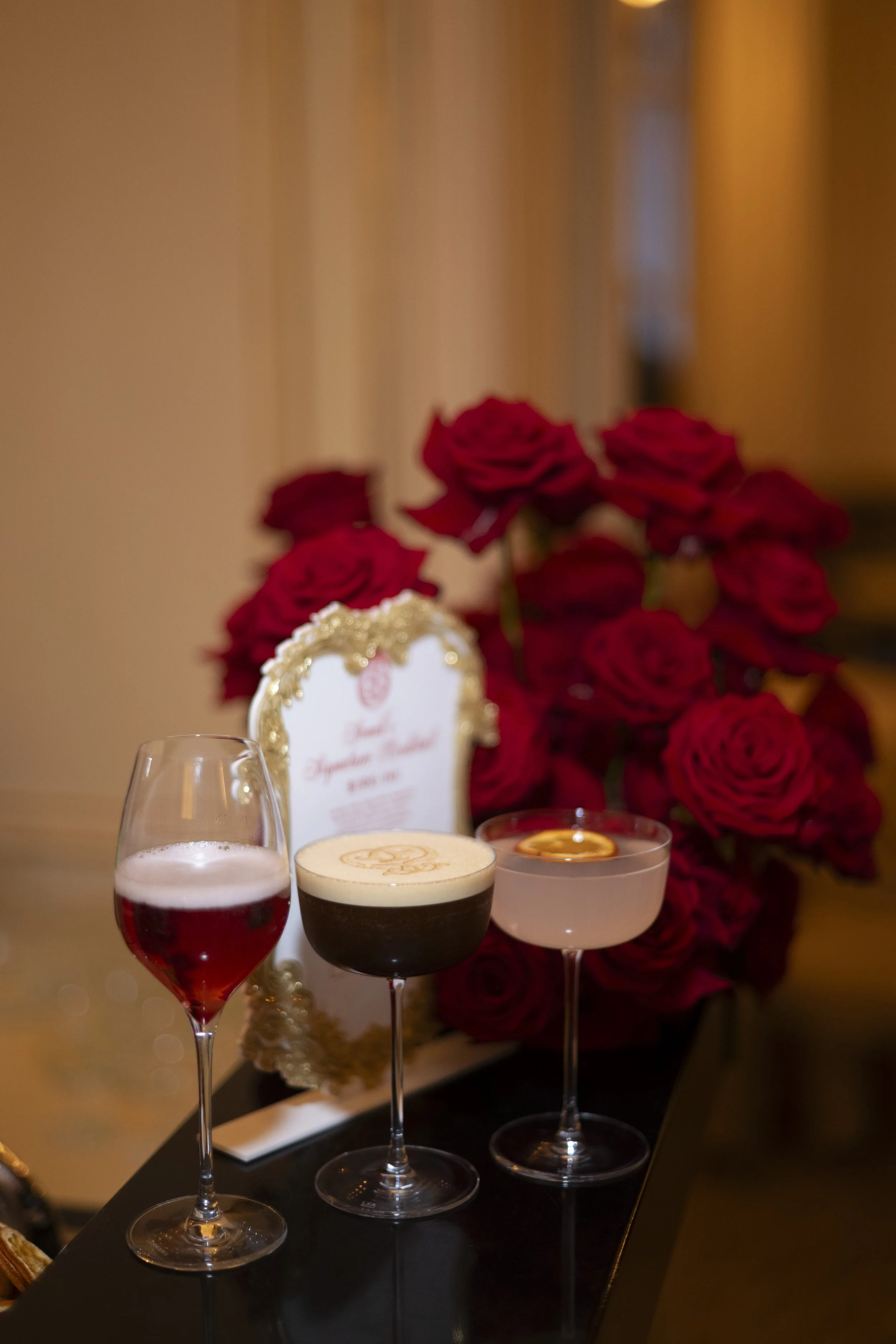 Four cocktails on a black tray with a bouquet of red roses and a decorative framed sign in the background.