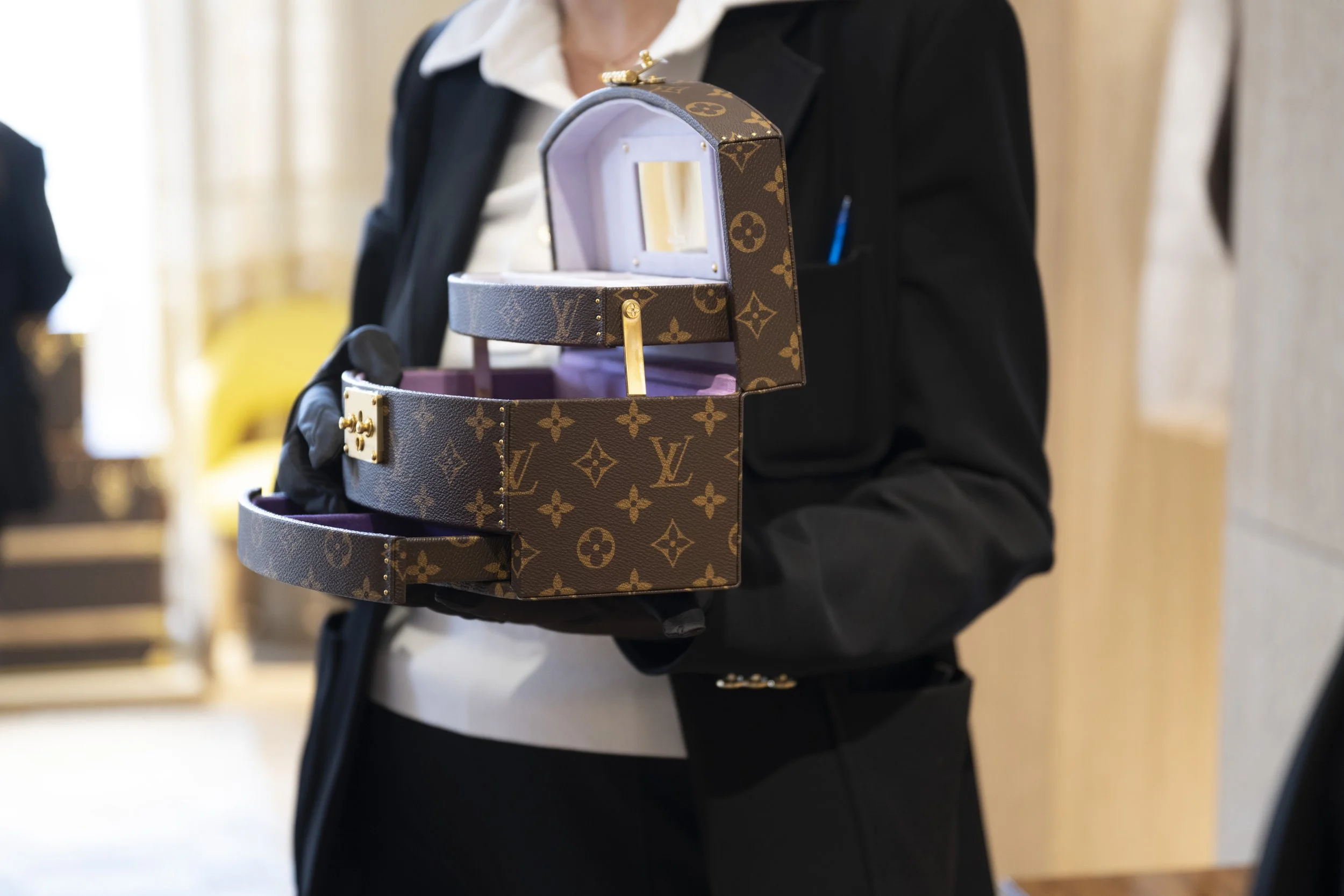 A person dressed in formal black attire holding a Louis Vuitton designer handbag shaped like a small shoebox, with the box open to reveal a glass display inside. The person's face is not visible.