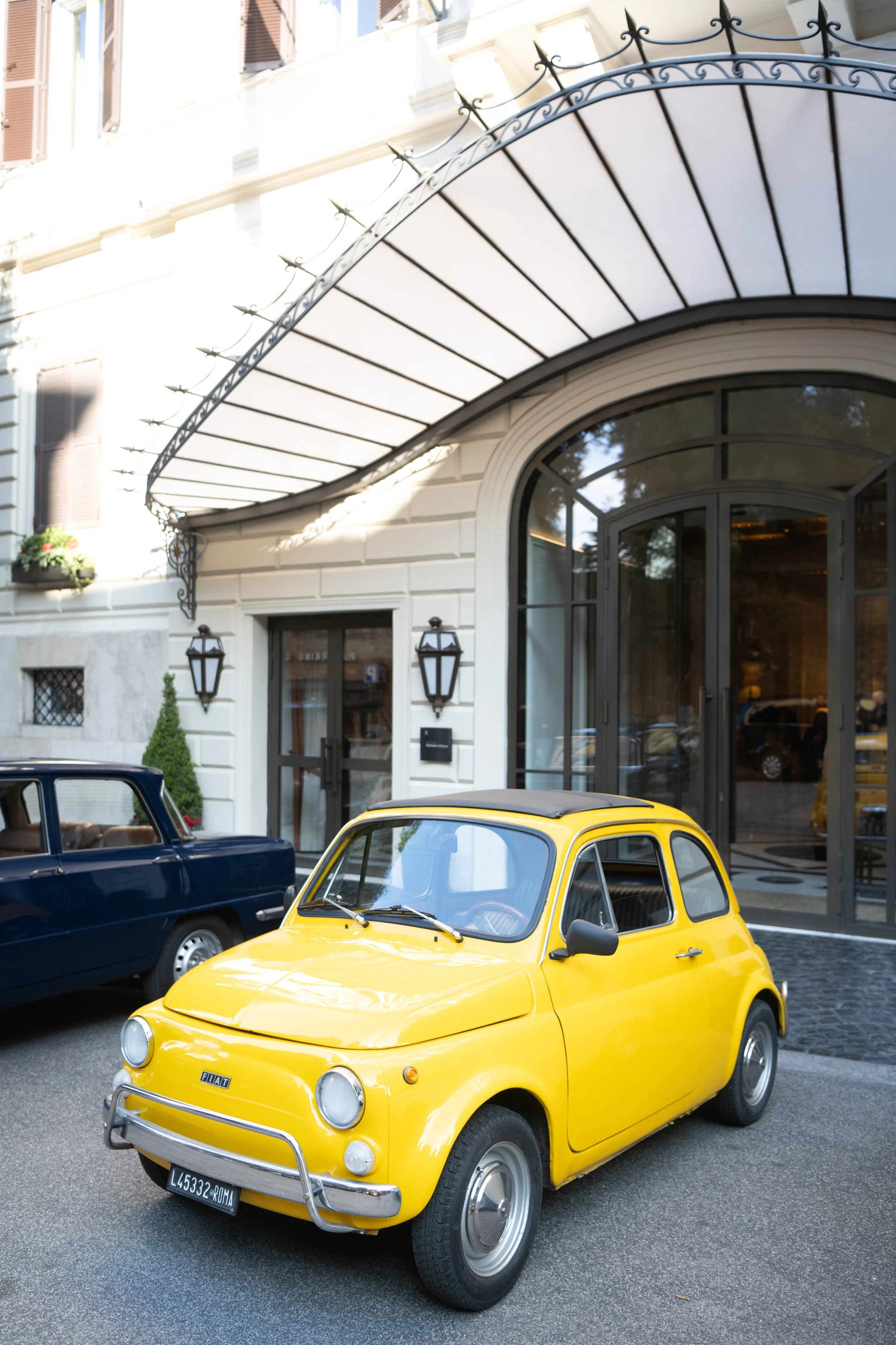 A bright yellow Fiat 500 parked on a city street, with a dark blue car beside it and a black car partially visible. The background features a white building with large glass doors, black lantern-style lights, and a decorative metal awning above the e