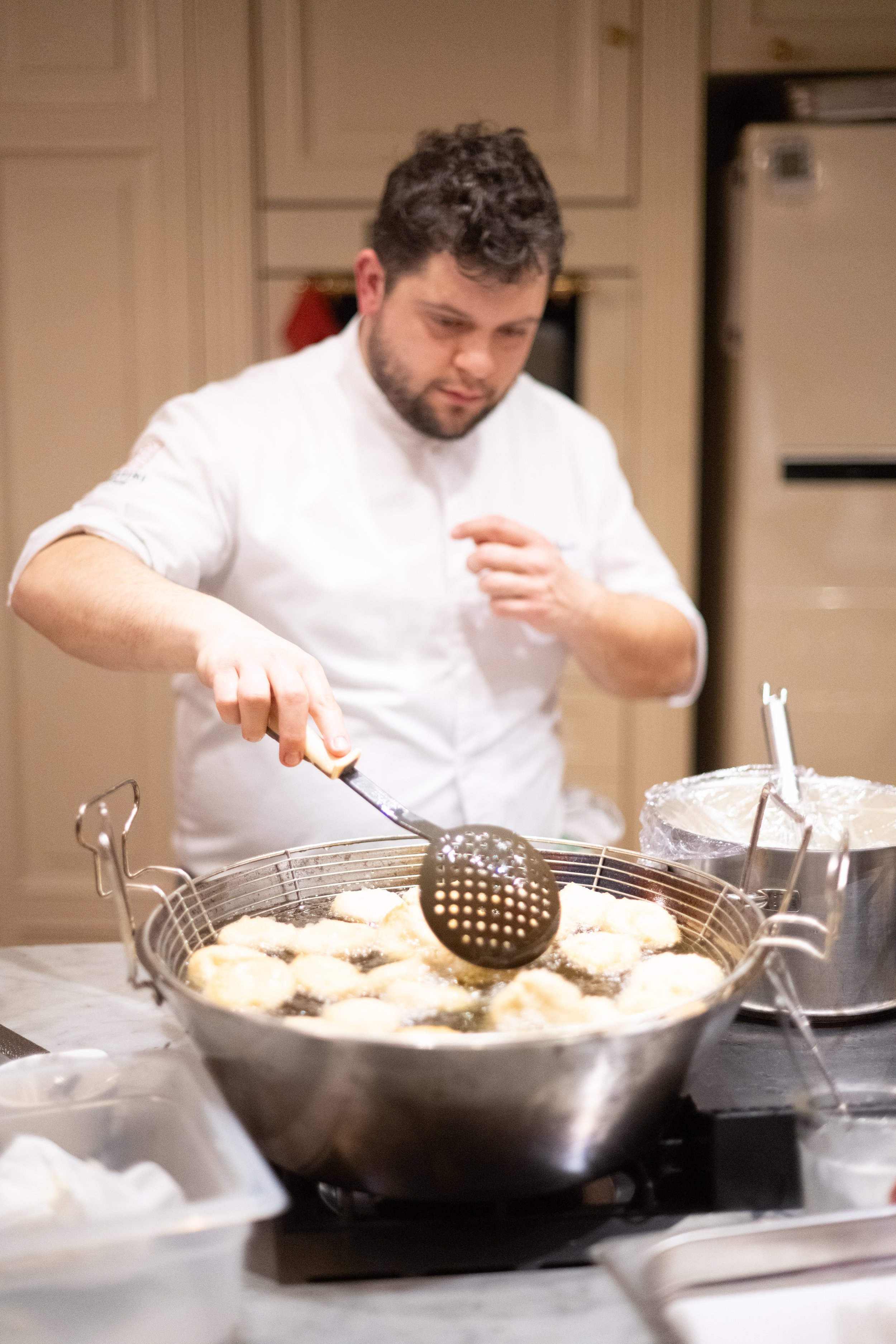A man in a white shirt is frying food in a large pan on a stove. The pan contains what appears to be multiple pieces of breaded food, possibly chicken or fish, with a slotted utensil in his hand. There are various kitchen items around him, including 
