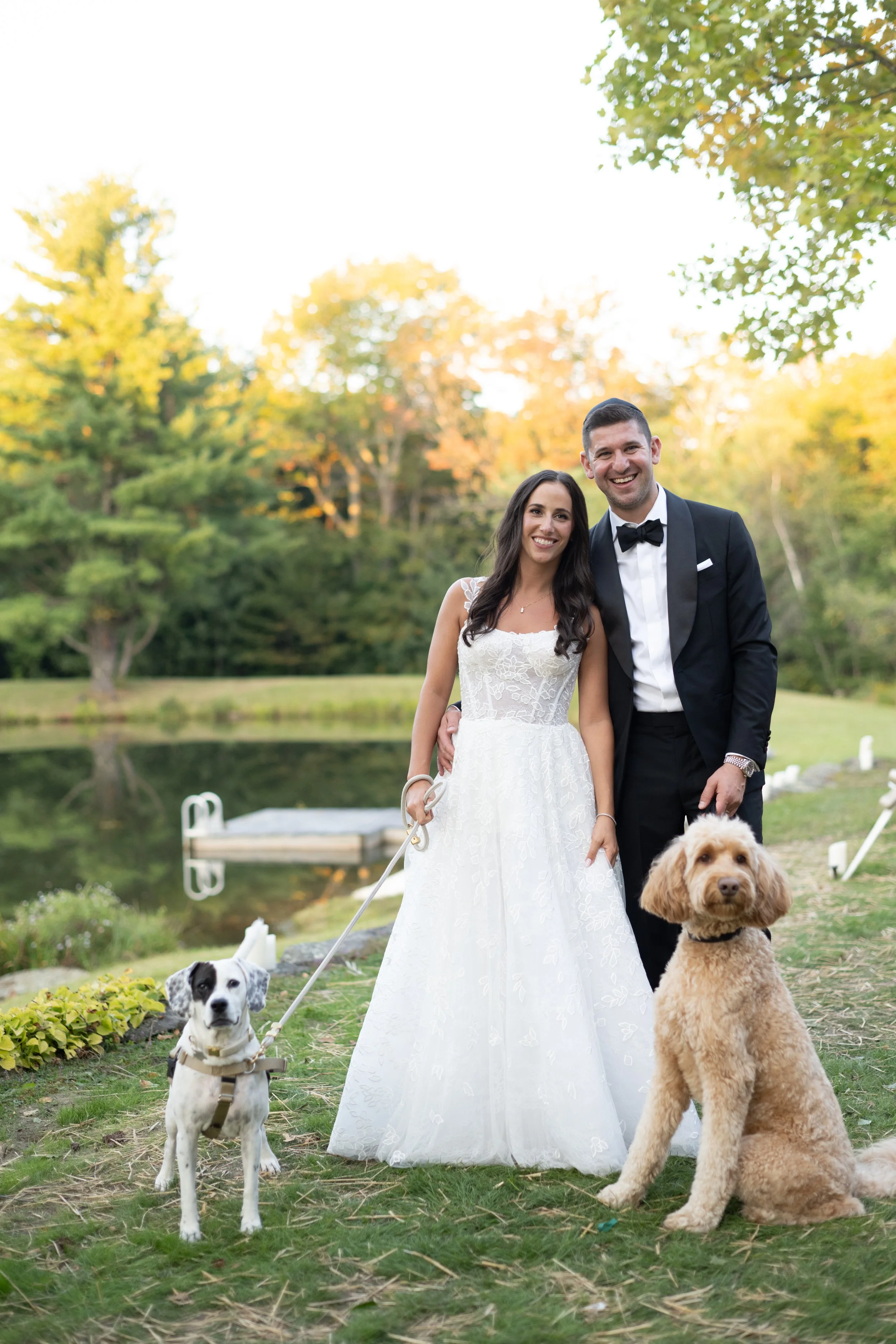 A smiling couple dressed in wedding attire standing outdoors with two dogs, a small black and white dog on a leash and a larger tan dog sitting on the grass. The background includes green trees with autumn leaves and a pond.