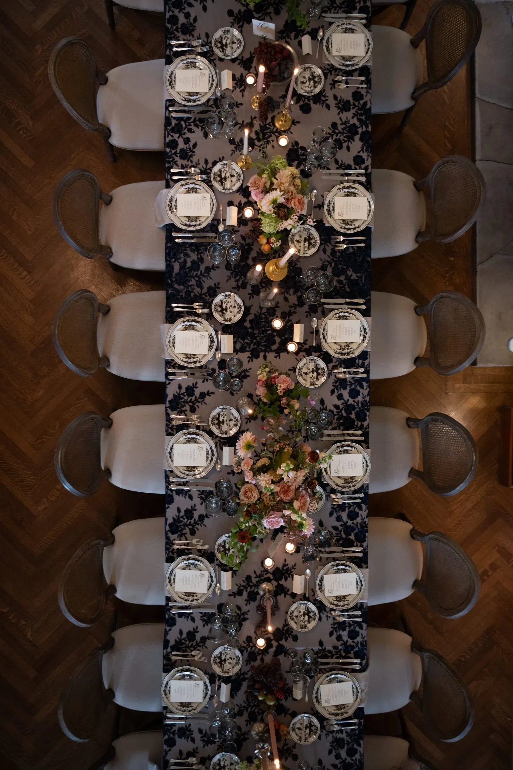 Overhead view of a elegantly set dining table with floral centerpiece, candles, and black and white patterned dishes, surrounded by cream-colored chairs.