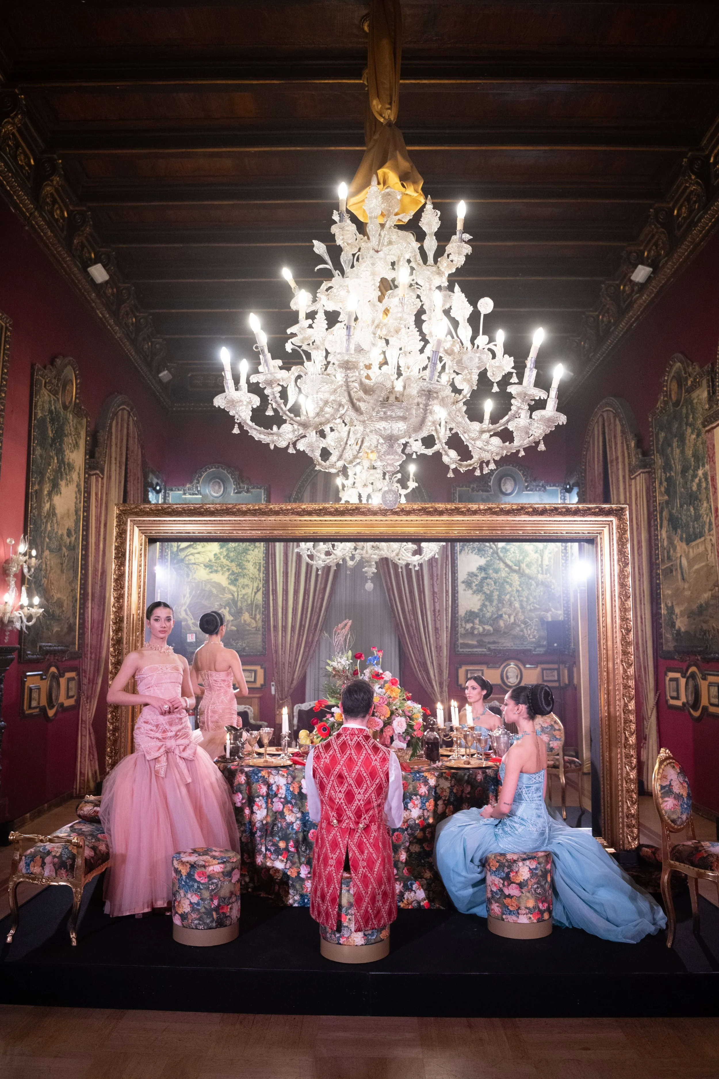 Elegant dining room scene with a large floral centerpiece, ornate chandelier, waitstaff in vintage attire, and guests in formal dresses, set against richly decorated walls with paintings and drapery.