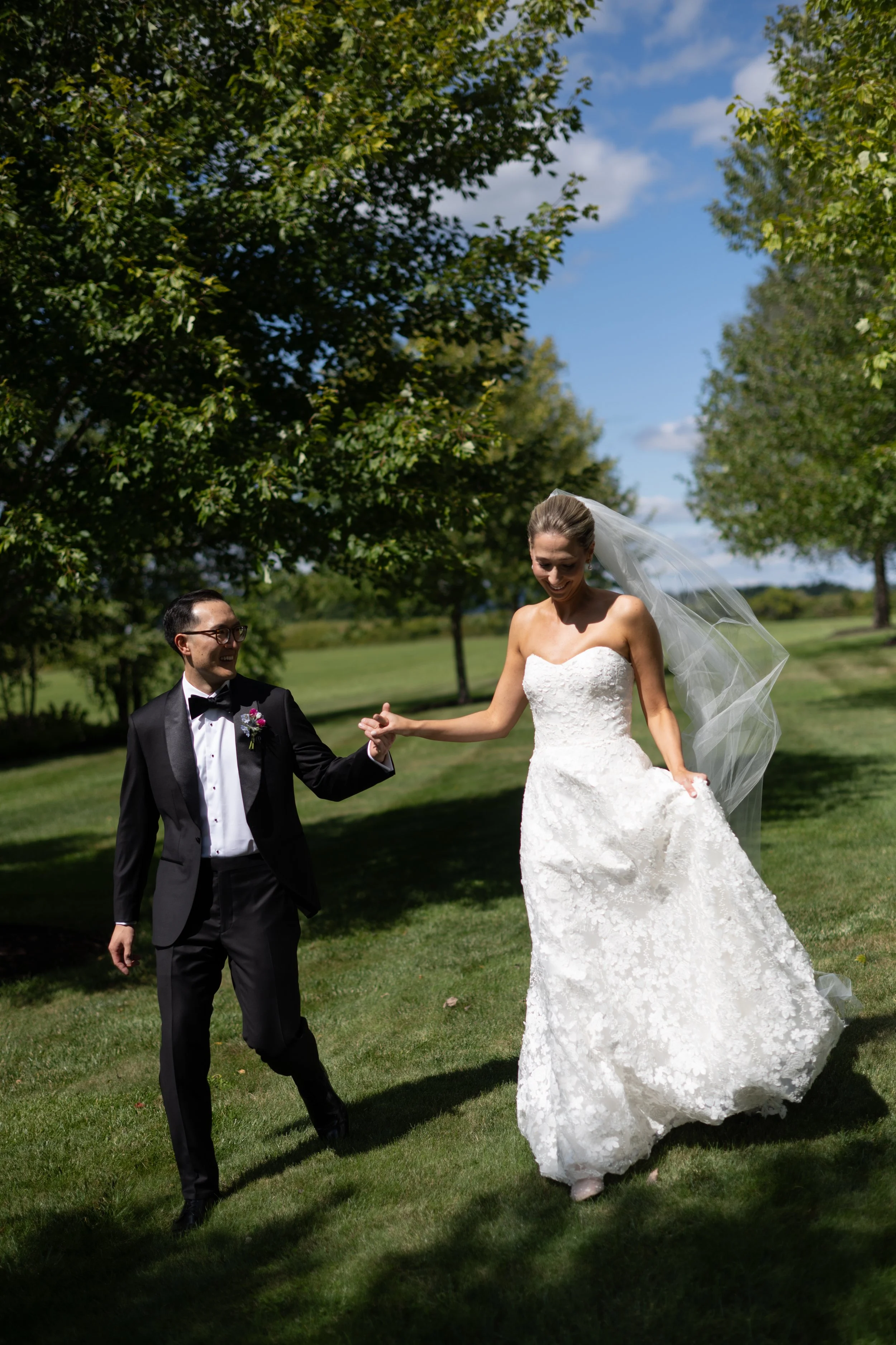 A bride in a white wedding dress with a veil and a groom in a black tuxedo with a bow tie and glasses are dancing together on a grassy field with trees and a blue sky in the background.