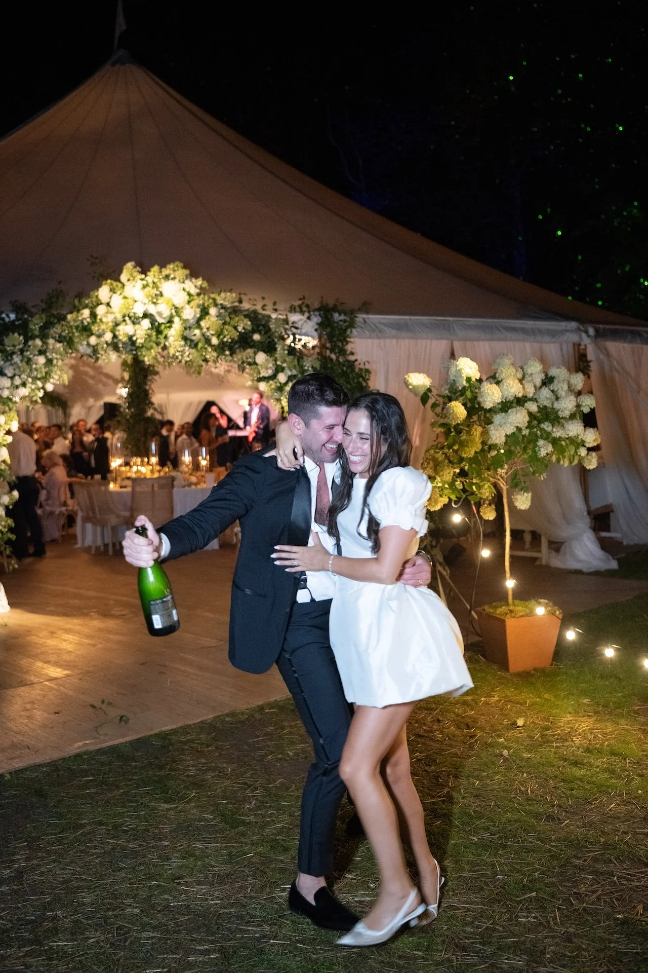 A couple dancing and smiling at a celebration under a large tent decorated with white flowers and string lights at night.