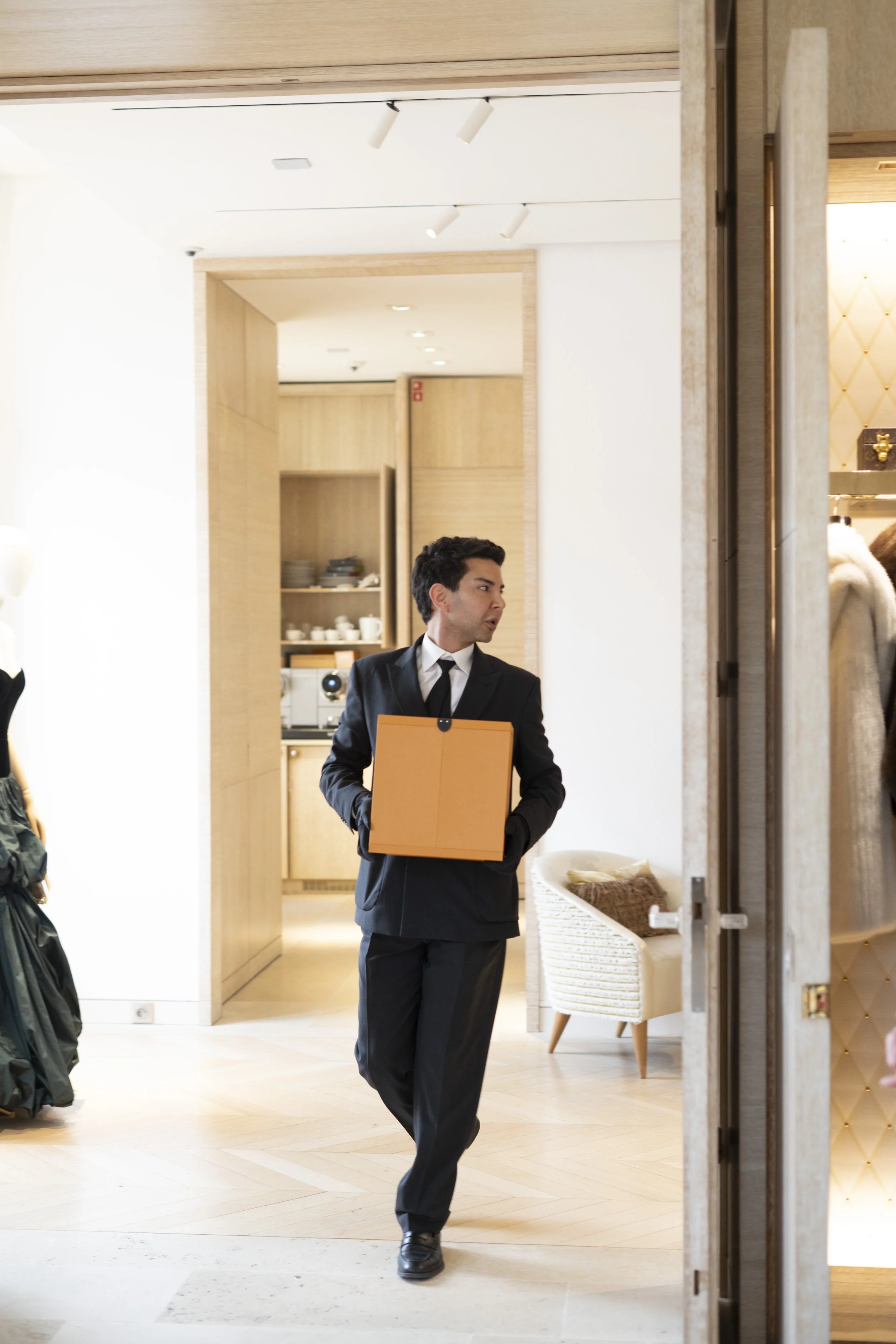 A man in a black suit and tie holding an orange folder is walking inside a modern, well-lit space with light wood paneling and furniture.