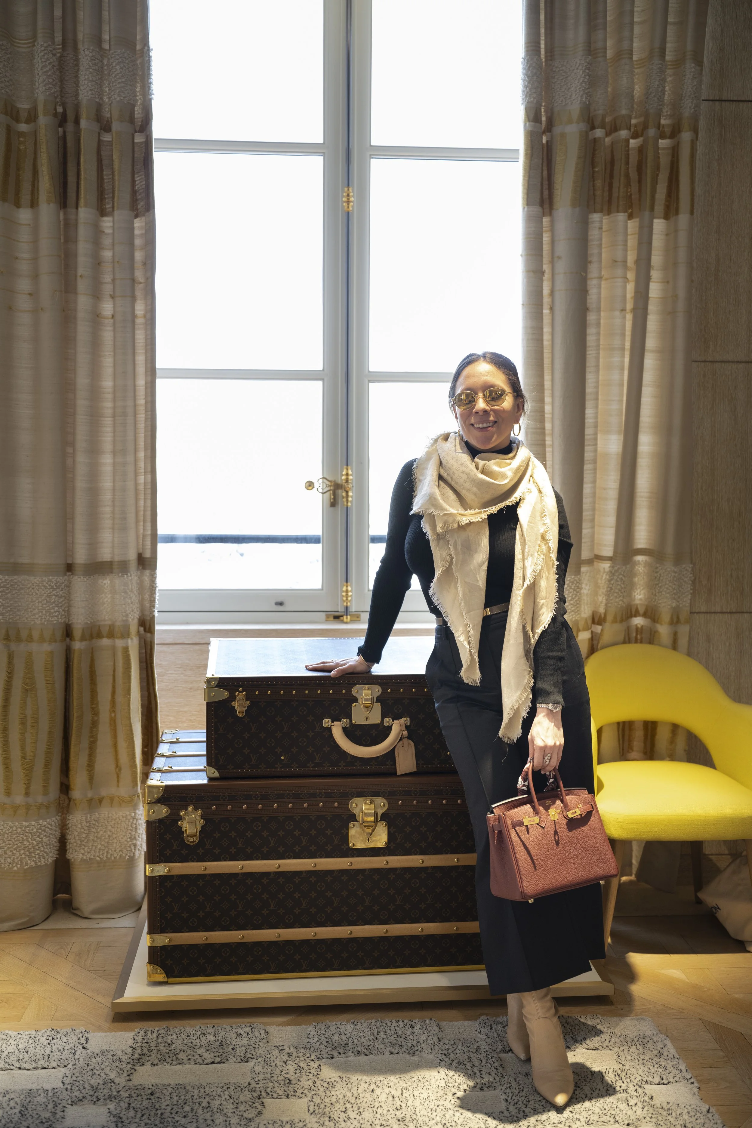 A woman wearing sunglasses, a beige scarf, black top, and black pants, holding a pink handbag, standing in front of a window with beige curtains, with a stack of three Louis Vuitton suitcases on a table and a yellow chair nearby.