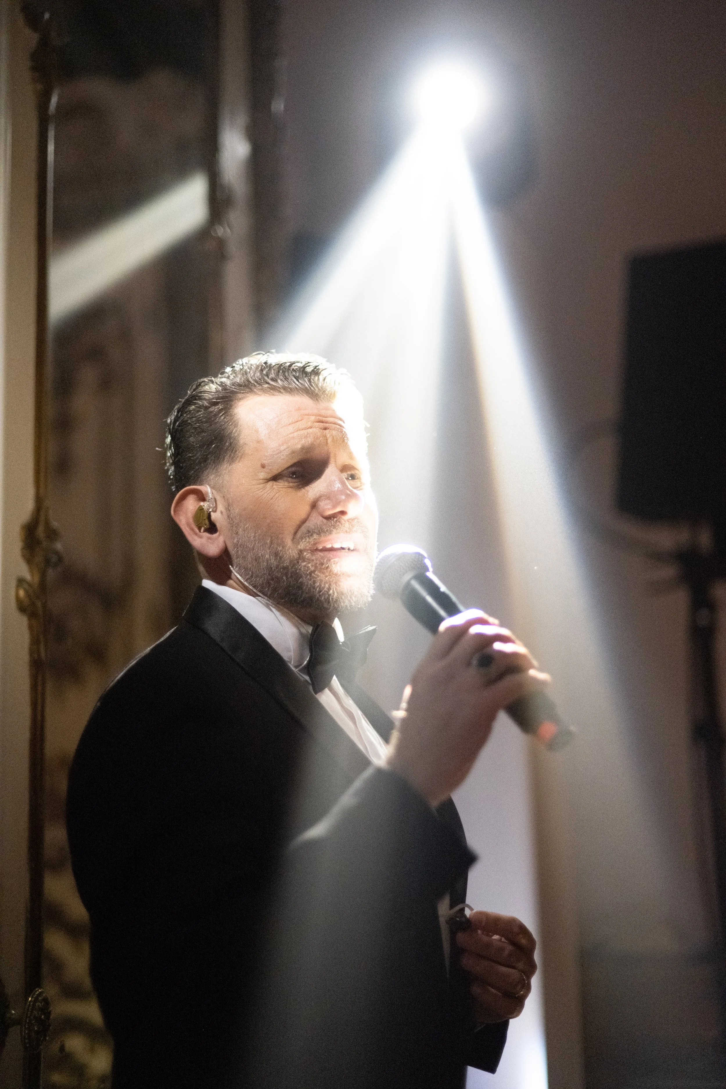 A man in a tuxedo holding a microphone and performing on stage, illuminated by a spotlight.