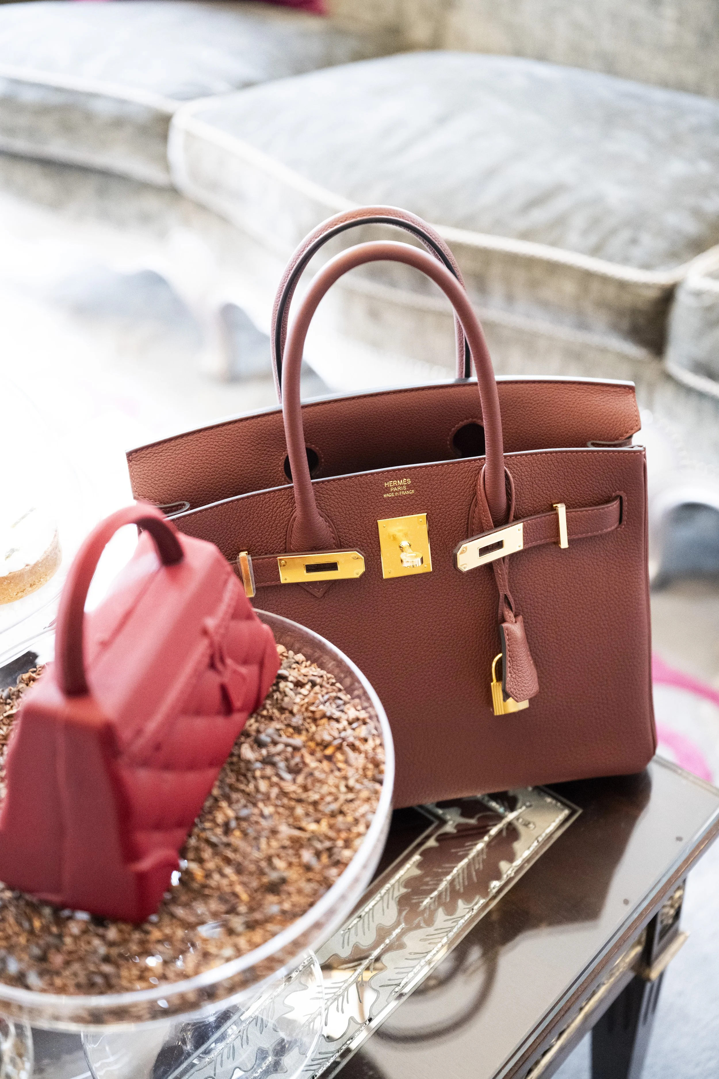 A close-up view of a pink Hermès handbag, a smaller pink handbag, and a tray with a bowl of granola. The handbag has gold hardware and is positioned on a reflective surface with a background of blurred trays and containers.