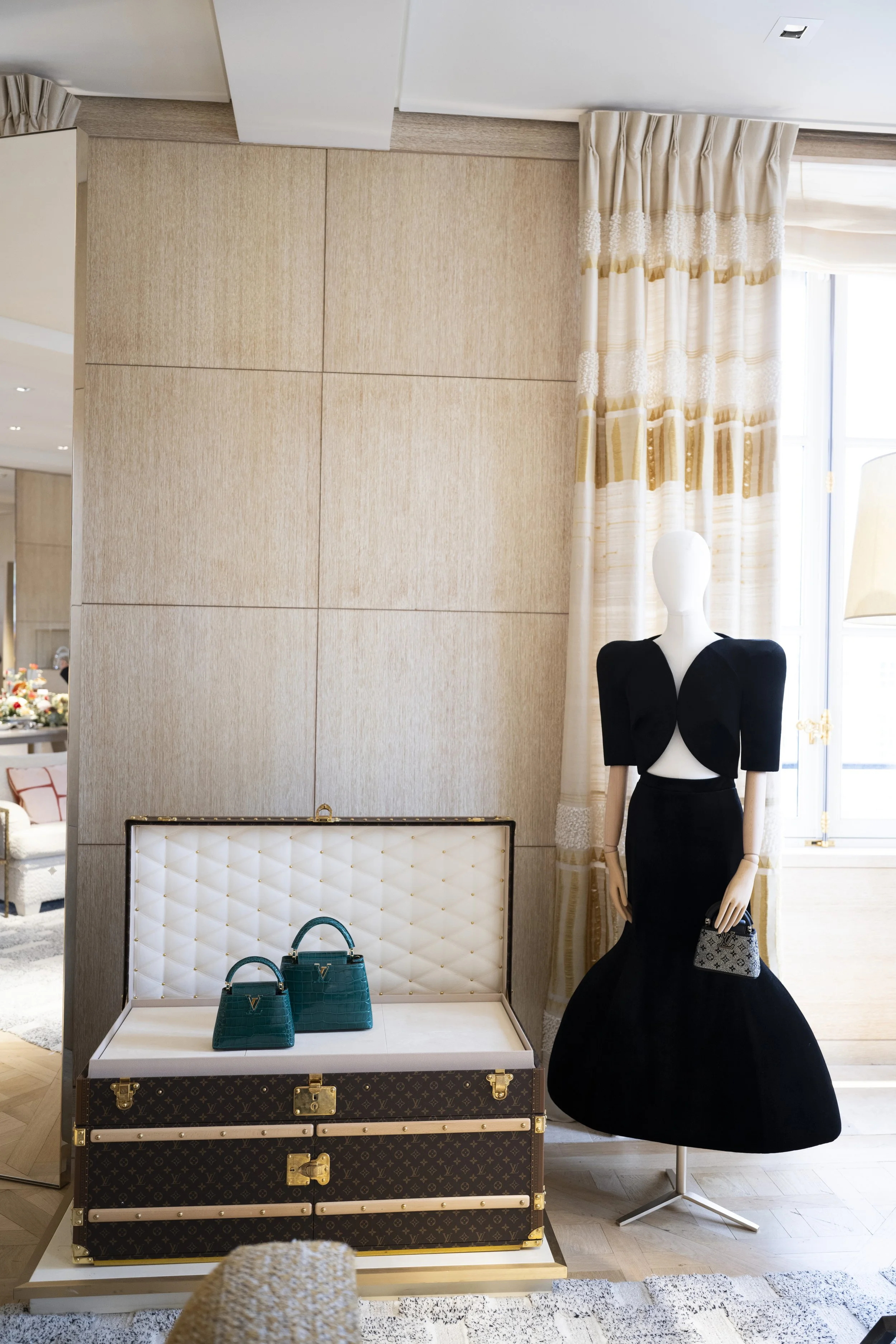 A mannequin dressed in a black dress with puffed sleeves and a fitted waist, holding a small black and white purse, standing next to a display table with two teal designer handbags. Behind them is a beige textured wall with vertical wooden accents, a