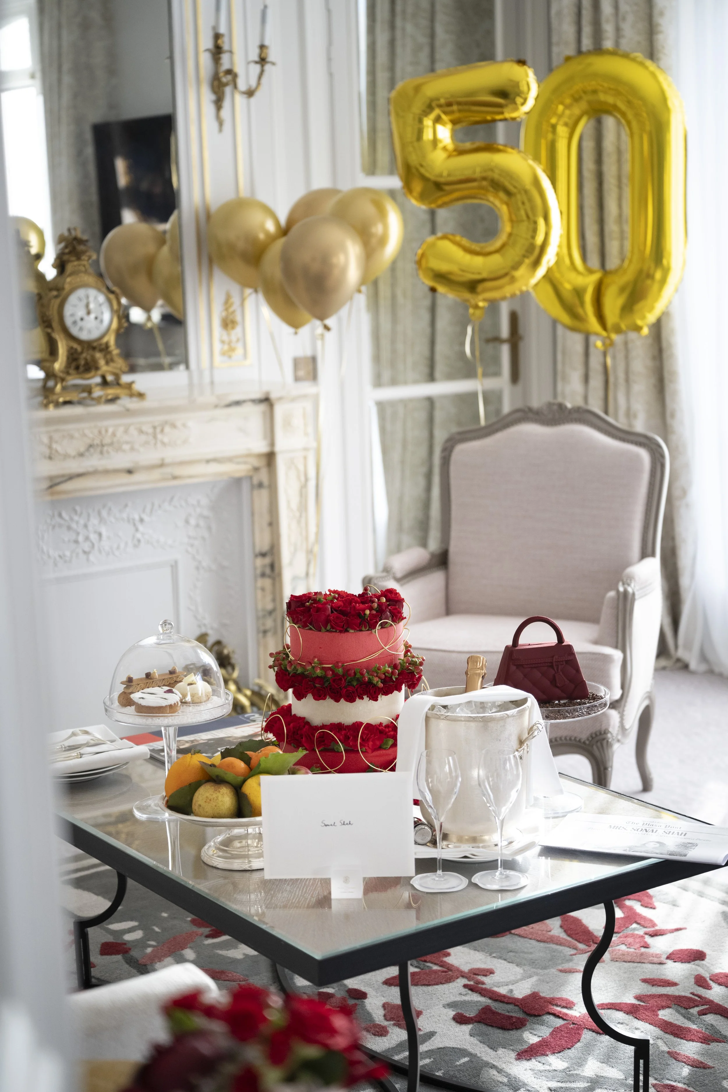 A decorated dining room with a glass table holding a red and white flower cake, fruit, and desserts. Gold balloons with the number 50 are hung above. Elegant chair with cream upholstery is in the background. Ornate antique clock and a mirror are on t