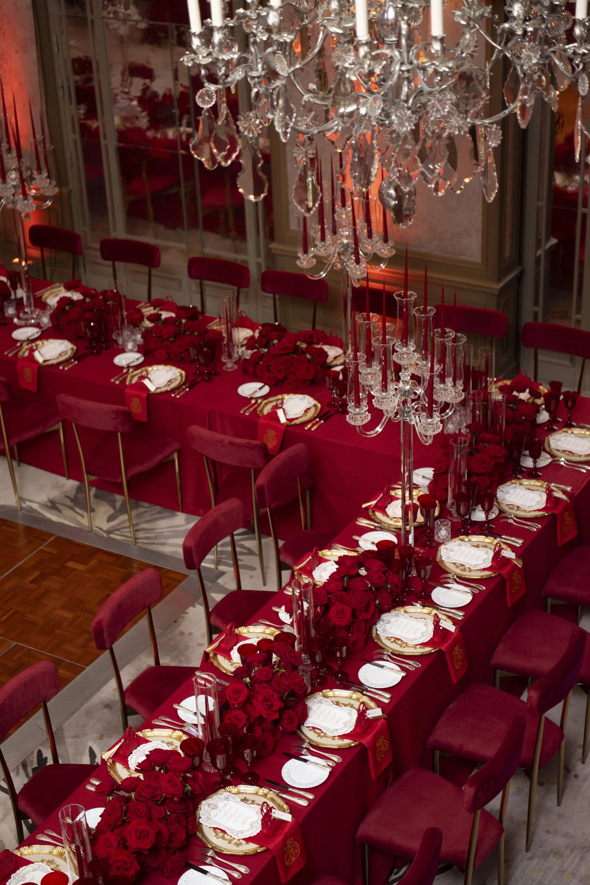 A luxurious dining table decorated with red roses, fine china, gold-rimmed plates, silverware, and tall red candles in glass holders, set in an elegant room with gold accents and a large crystal chandelier overhead.