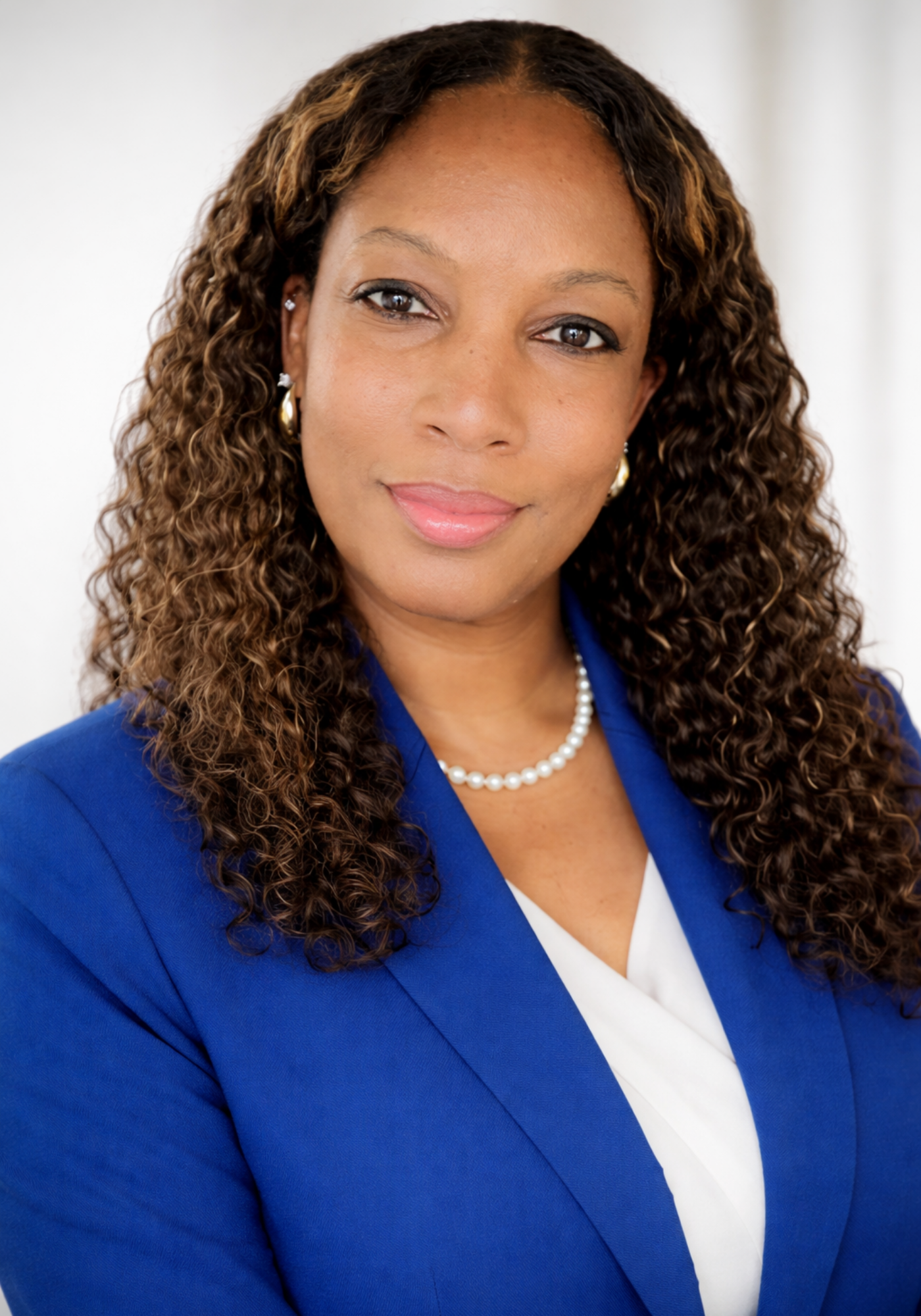 A woman with curly brown hair, wearing a blue blazer, white blouse, pearl necklace, and earrings, smiling softly at the camera.