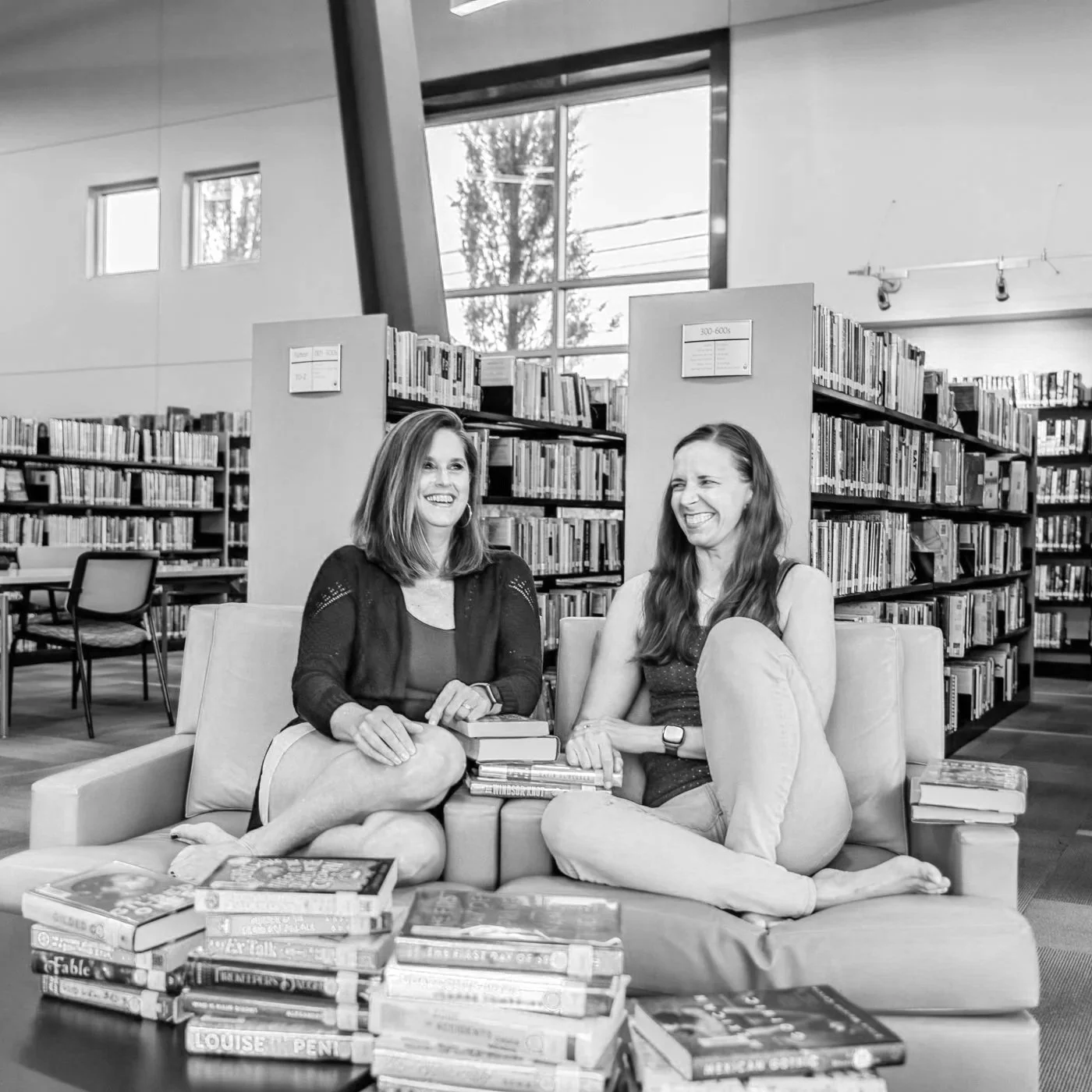 Meredith (left) and Kaytee (right) side on two adjoining chairs at the library. They are surrounded by library shelves and piles of books on the table in front of them. They are not looking at the camera but are laughing together.