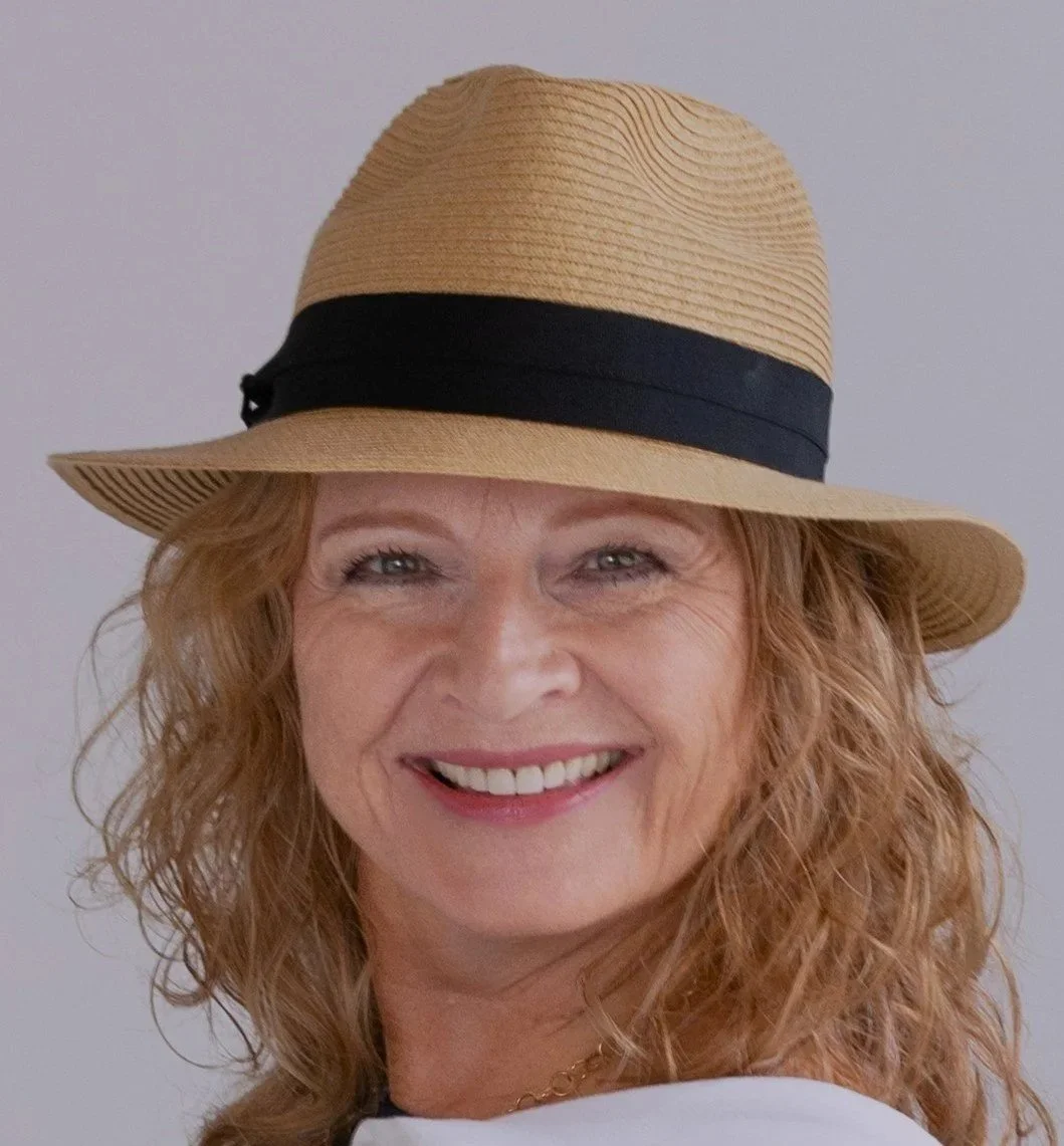 A woman with curly red hair wearing a tan wide-brimmed hat with a black band, smiling at the camera.