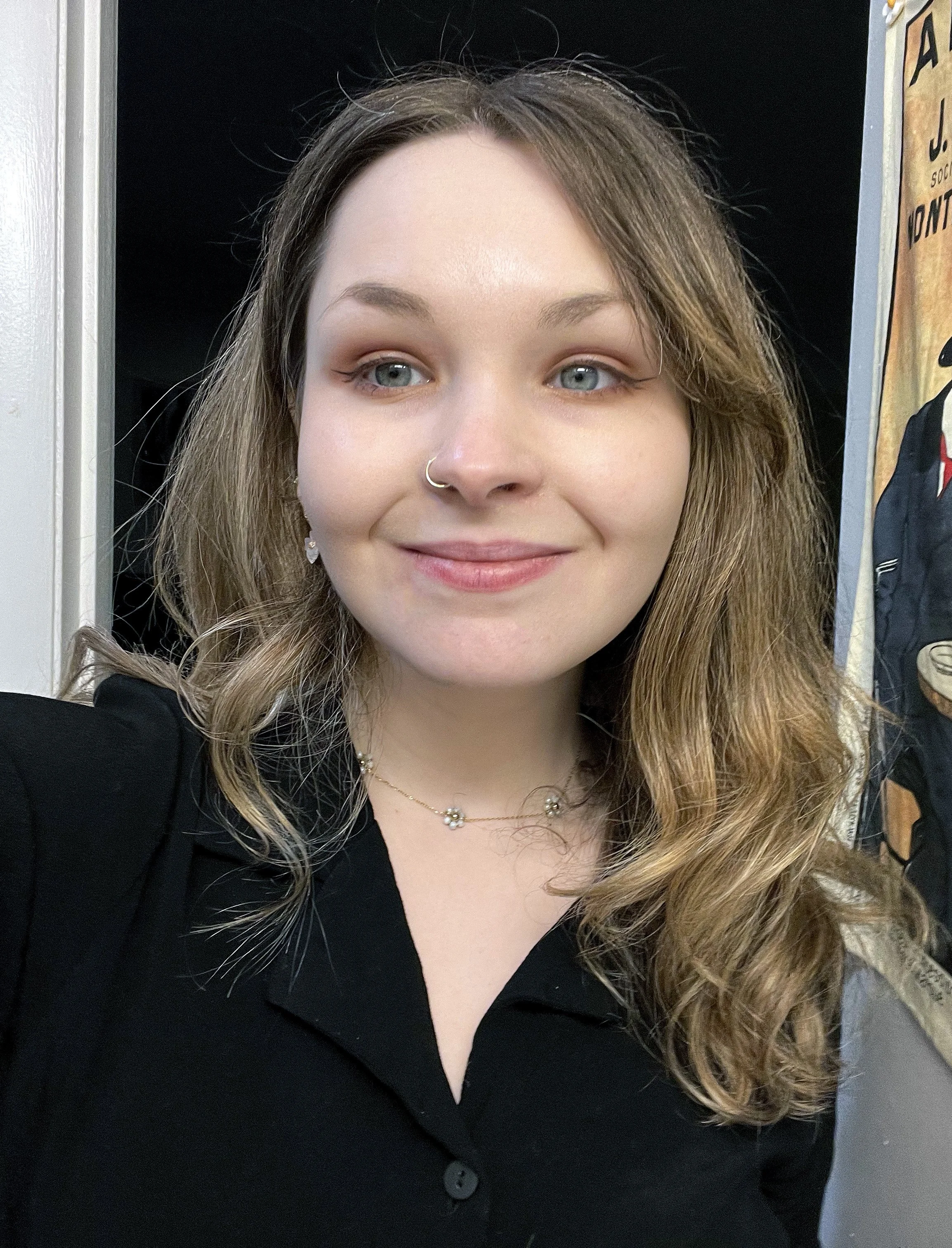 A young woman with light skin, shoulder-length wavy brown hair, and blue eyes, smiling, wearing a black top, a necklace with small flowers, and a nose ring, standing indoors near a doorway with posters on the wall to her right.