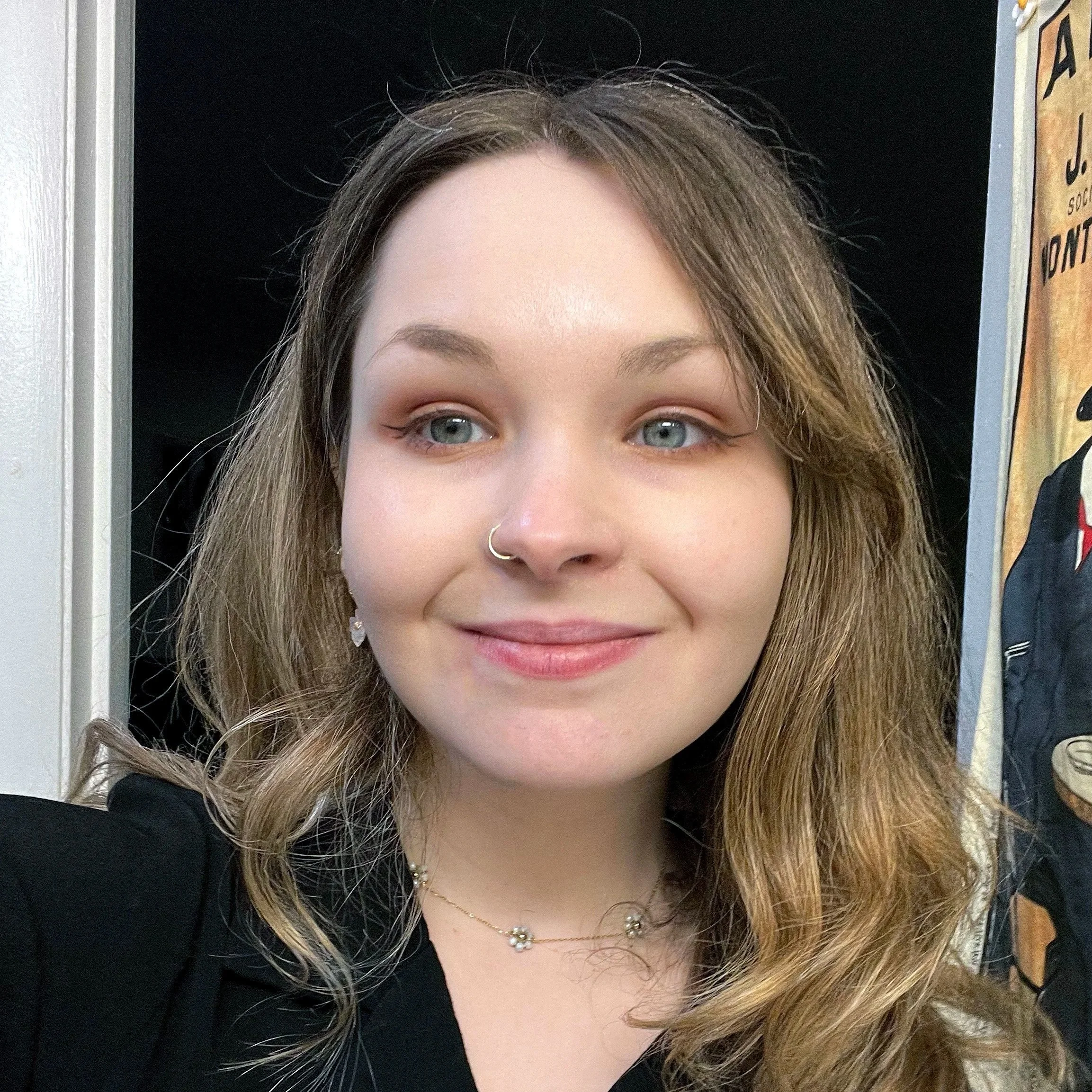 Close-up of a young woman with fair skin, wavy light brown hair, blue eyes, wearing a nose ring, earrings, and a delicate necklace, smiling at the camera.