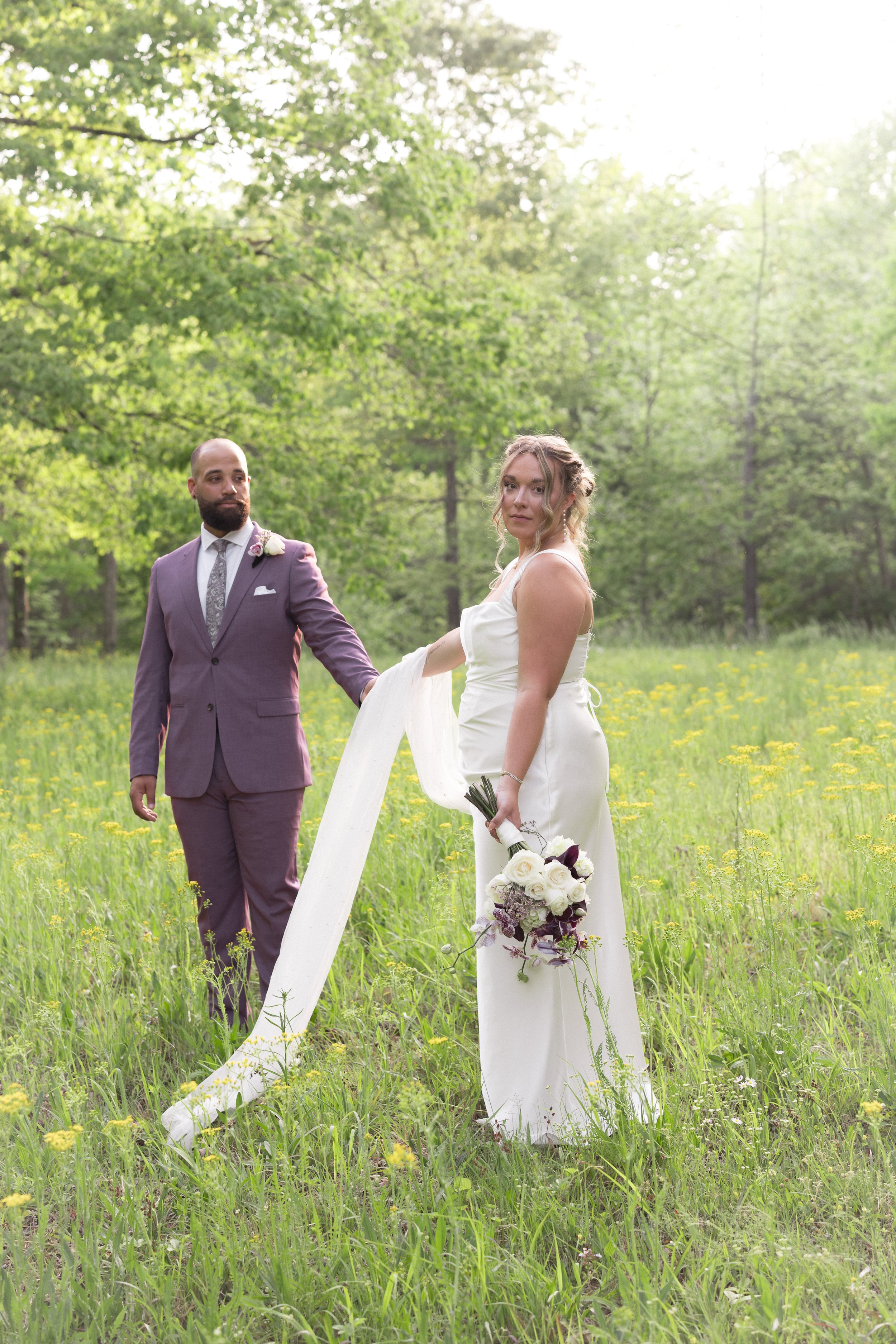 A bride and groom holding hands in a grassy field with yellow flowers, surrounded by green trees, with sunlight shining through the foliage.