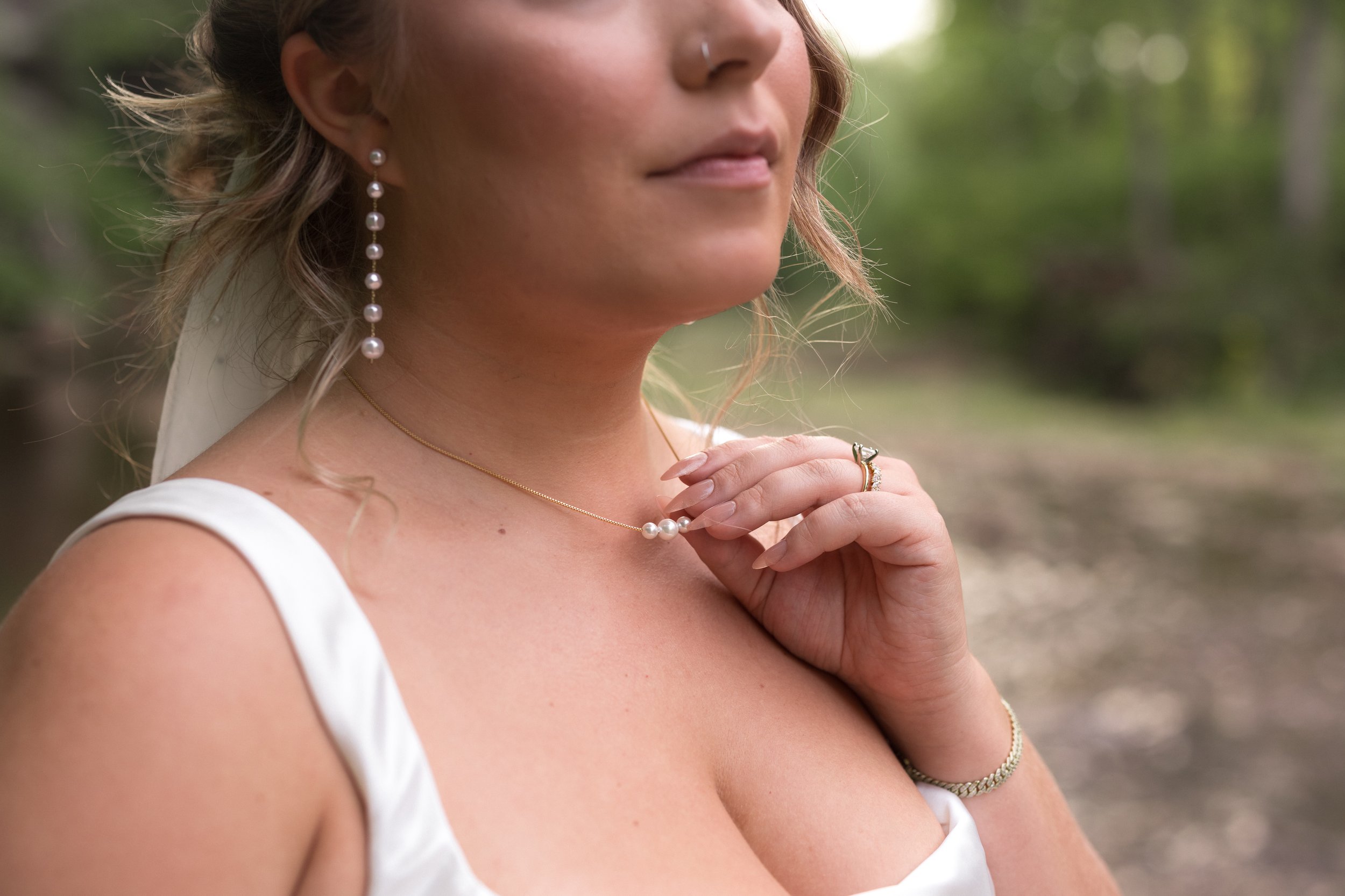 Close-up of a woman's neck and face with pearl earrings, a gold necklace, a ring, and a nose piercing, outdoors with blurred green background.