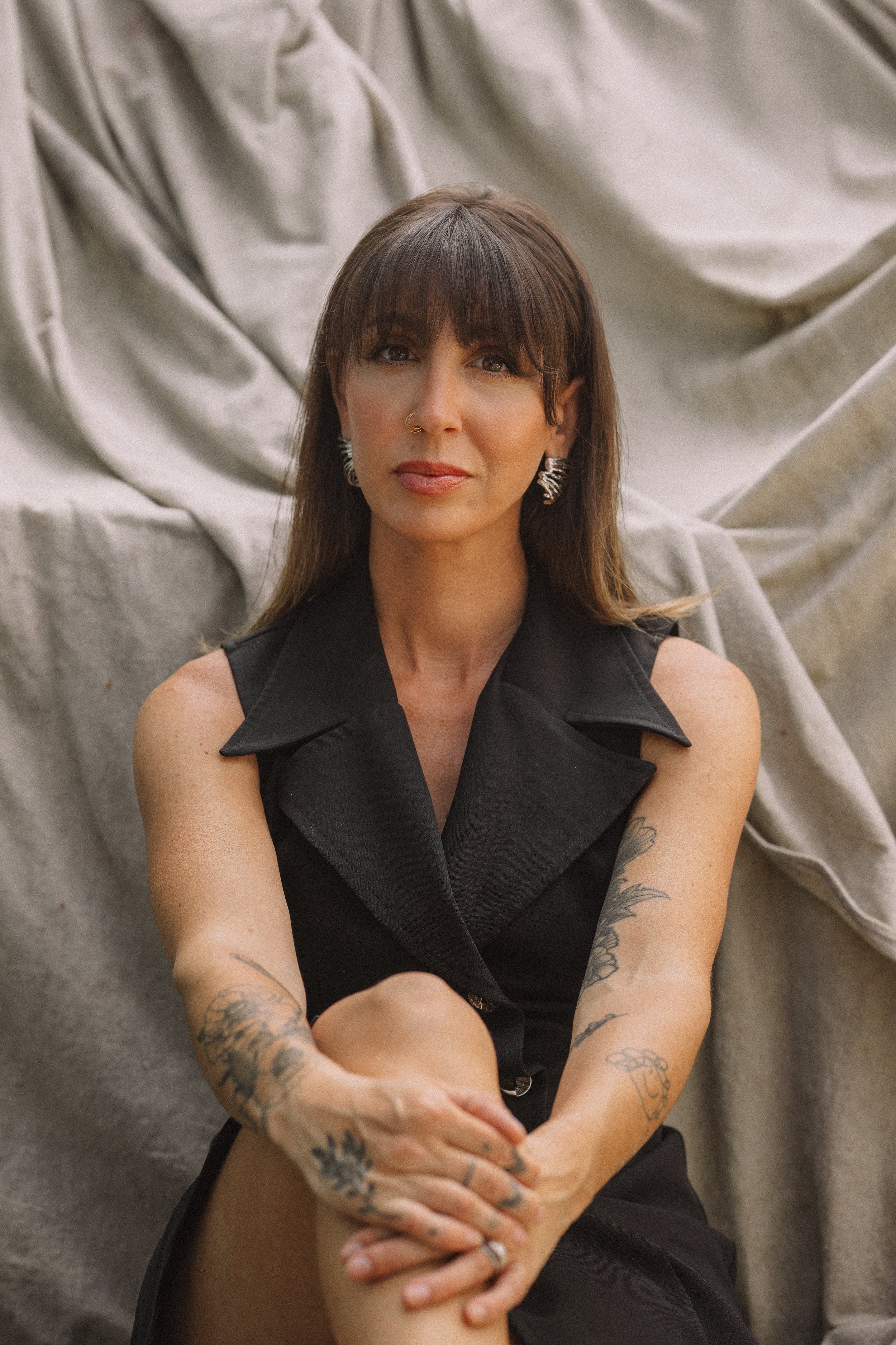A woman with long brown hair, wearing a black sleeveless top with a collar, sitting in front of a draped fabric backdrop. She has various tattoos on her arms and is wearing jewelry including earrings, a nose ring, and rings. She is looking directly at the camera with a neutral expression.