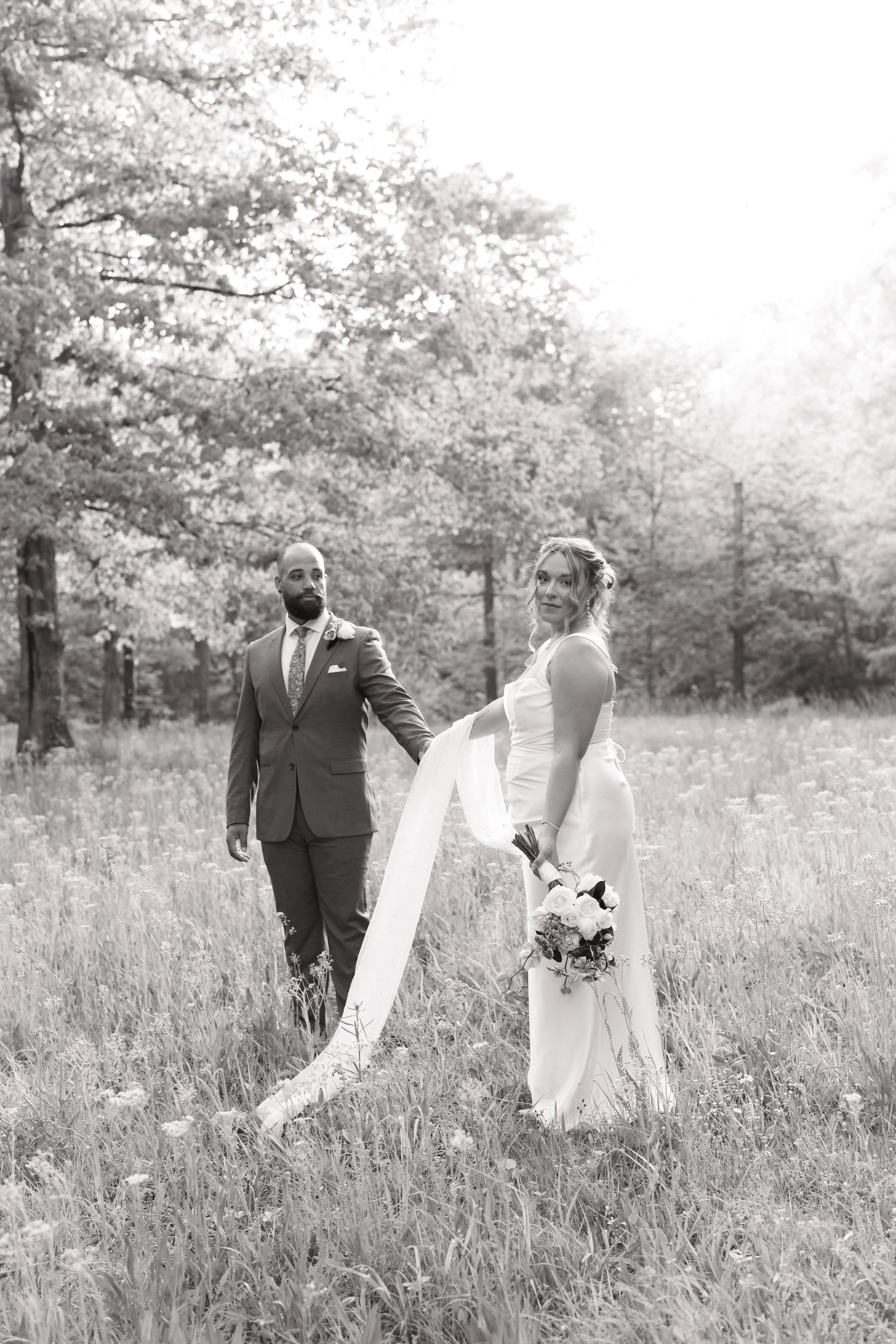 Black-and-white photo of a bride and groom holding hands in a field with tall grass and trees in the background.