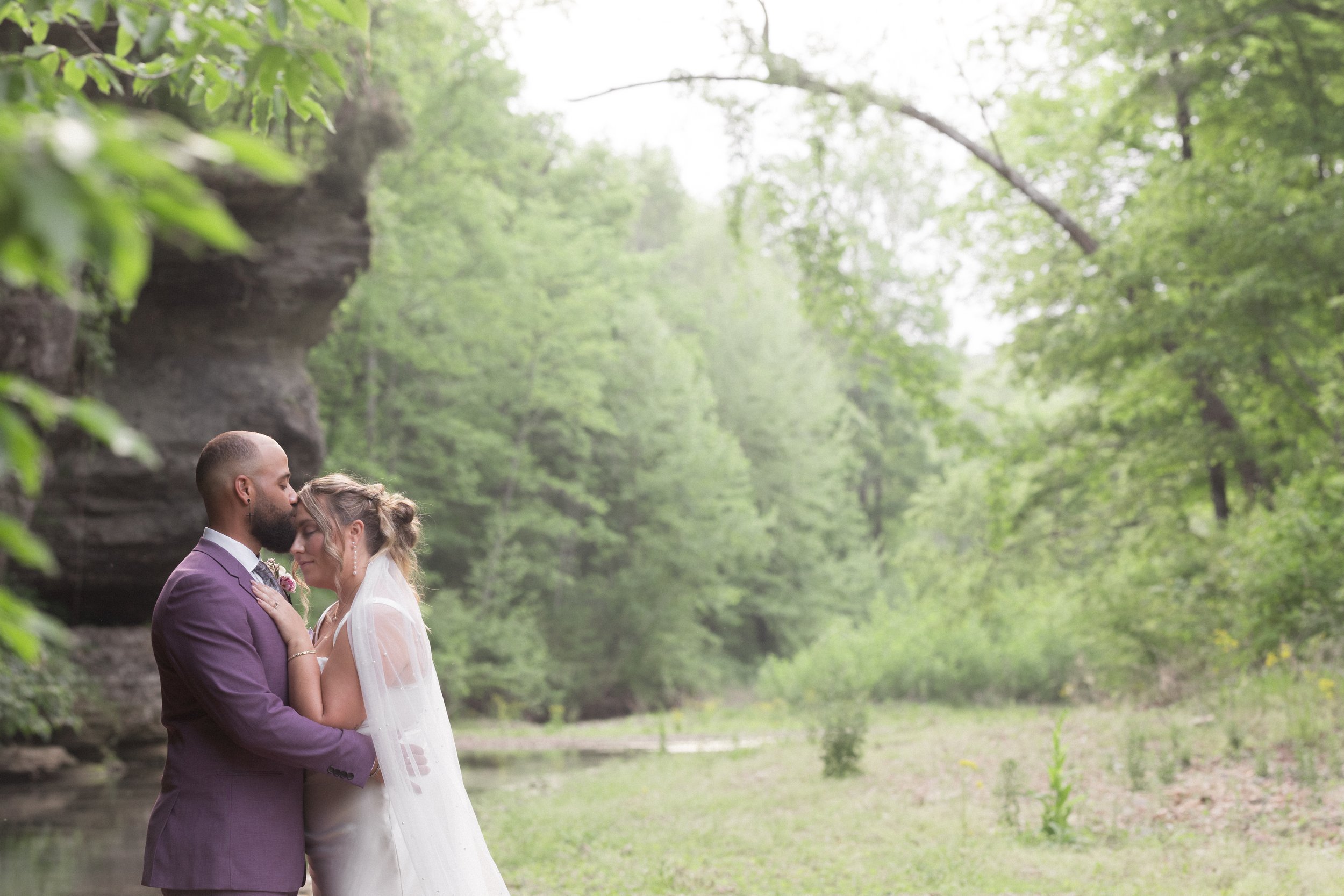 A couple in wedding attire sharing a kiss and embracing outdoors in a lush green, wooded area.