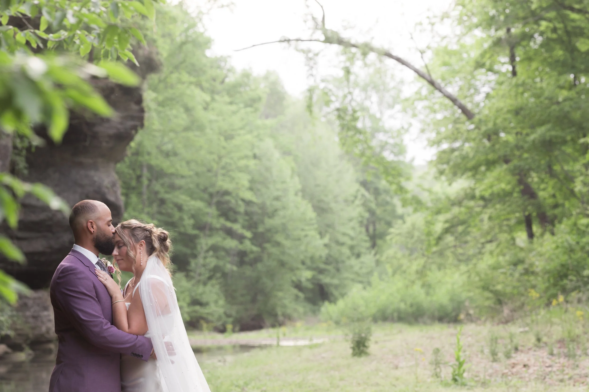 A couple dressed in wedding attire sharing an intimate moment outdoors in a lush, green forest.