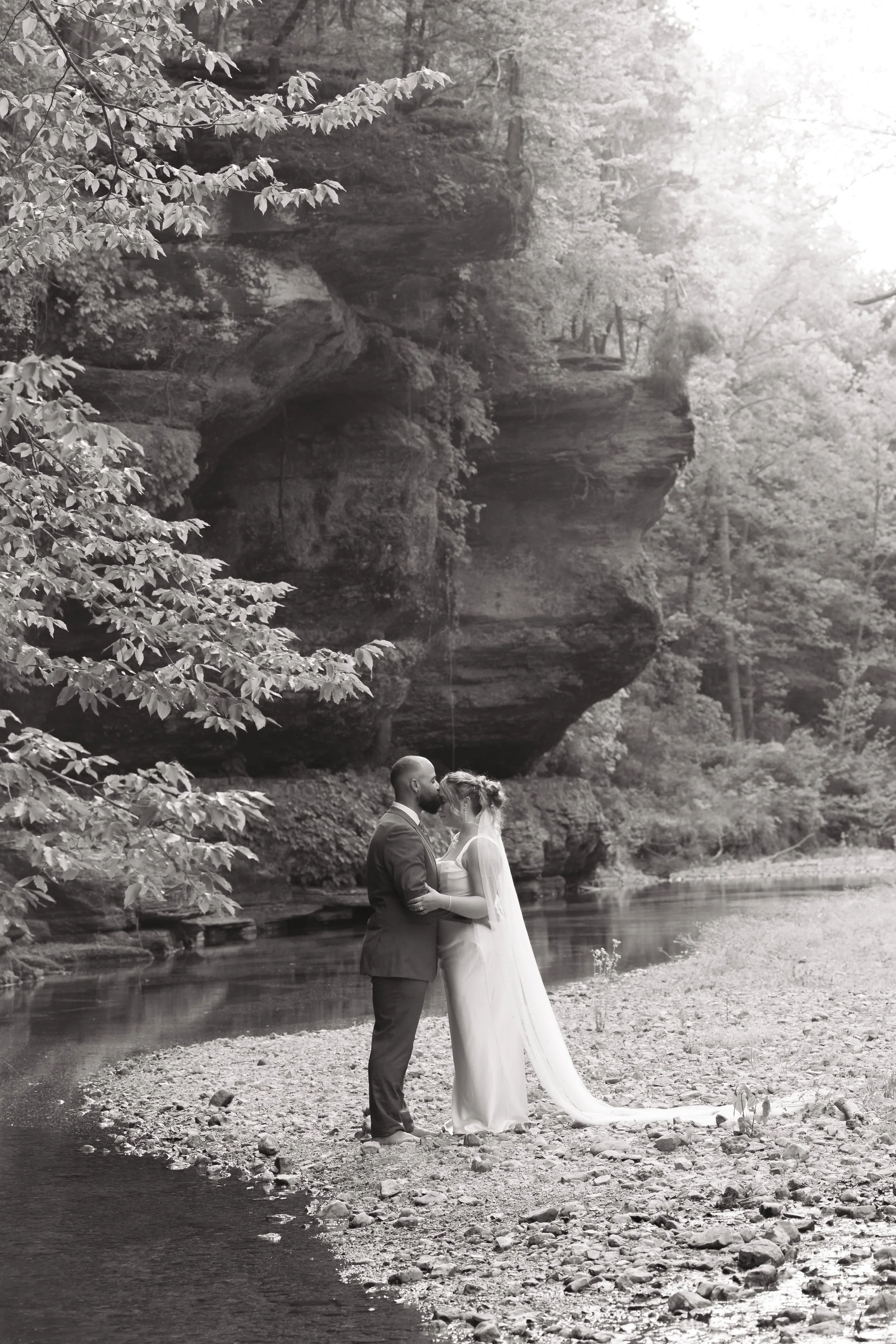 A black and white photo of a bride and groom standing on the rocky shore of a river, with tall trees and a large rock formation in the background. The couple is holding each other and looking into each other's eyes.