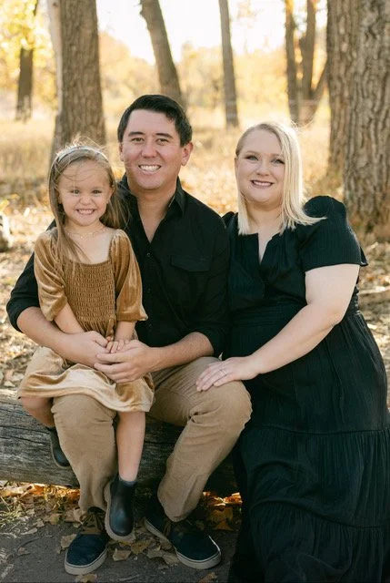Family of three sitting outdoors in a forest during autumn, smiling at the camera.