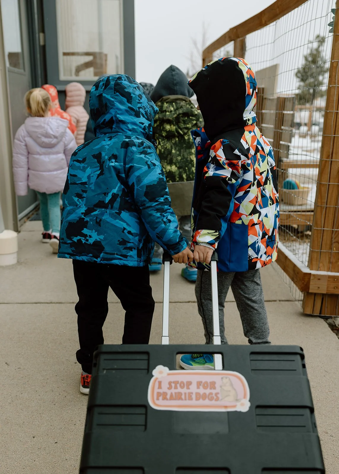 Two children wearing winter jackets and hoodies, pulling rolling suitcases, stand facing each other outdoors near a wire fence. In the foreground, a black case with a sticker reading 'I STOP FOR PRAIRIE DOGS' is visible.