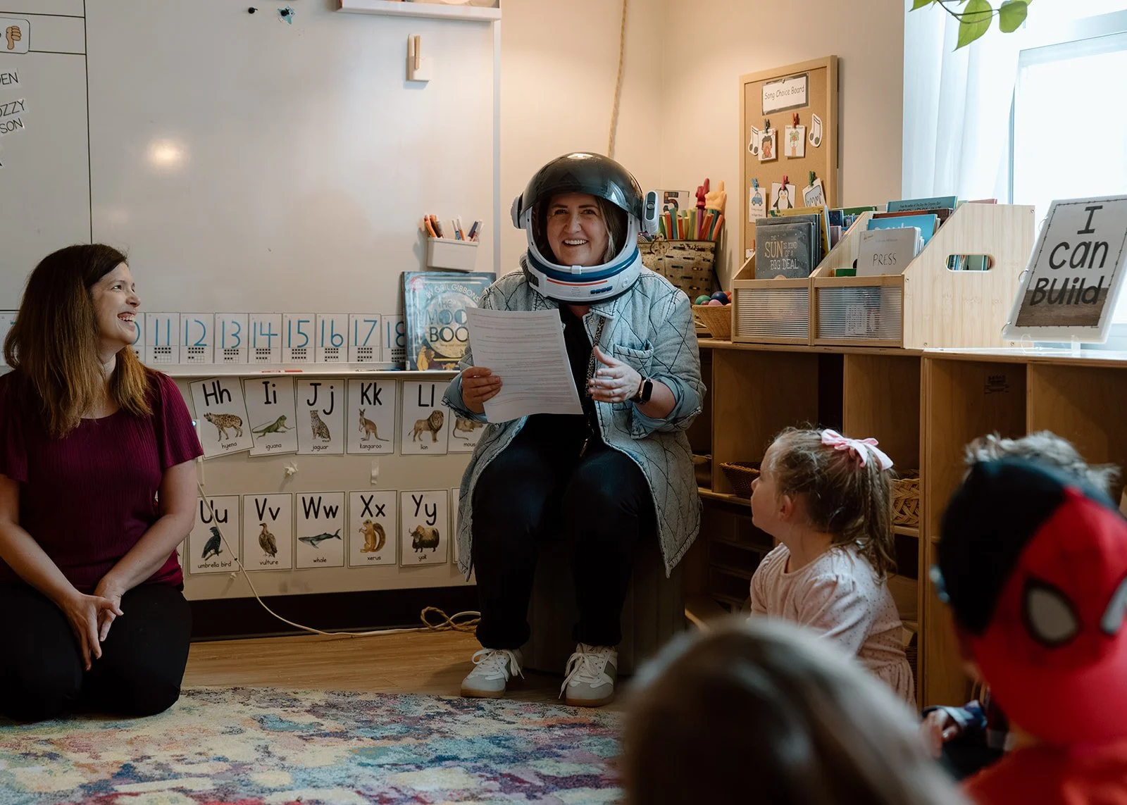 A woman sitting on a chair wearing a space helmet, holding a paper, in a classroom with young children and another woman sitting on the floor nearby. The classroom has educational posters and books.