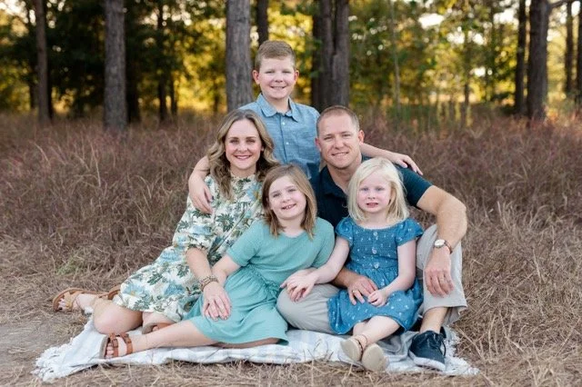 Emily Arnold family photo with 3 children and husband in a field with trees in the background.