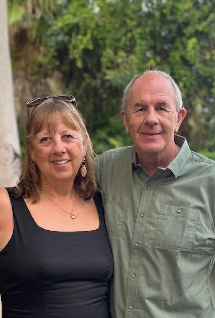 A smiling middle-aged woman and man standing outdoors in front of green trees, with the woman wearing a black top and the man wearing a green button-up shirt.