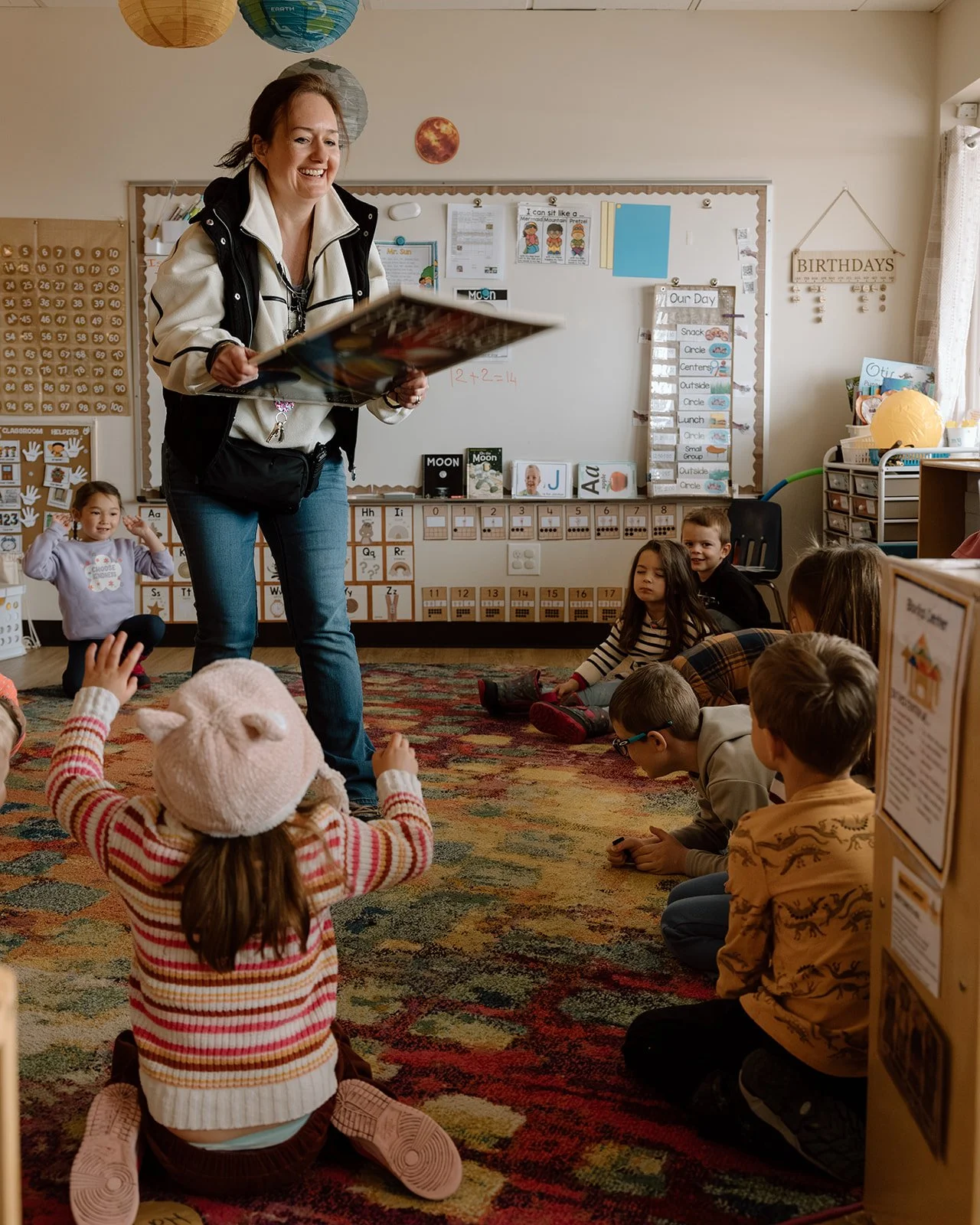 A classroom with a teacher reading a book to young students sitting on the colorful carpet. The students are facing the teacher, some raising their hands. The room is decorated with educational posters, a whiteboard, and colorful hanging lanterns.