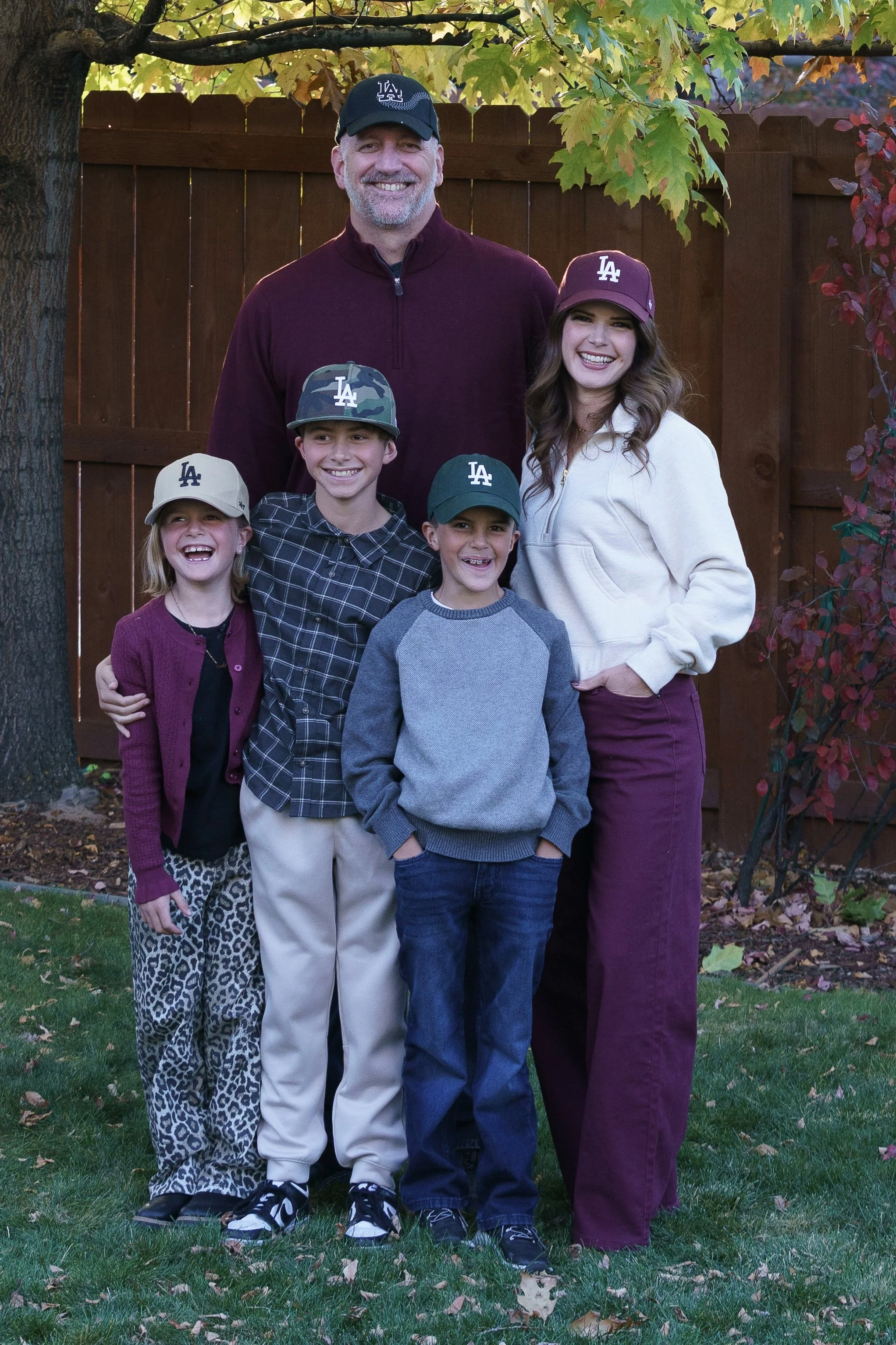 Family of five standing outdoors in front of a wooden fence with autumn leaves on the ground and a tree with colorful leaves overhead. They are all wearing casual clothing with some family members wearing Los Angeles Dodgers baseball caps. The adults and children are smiling.