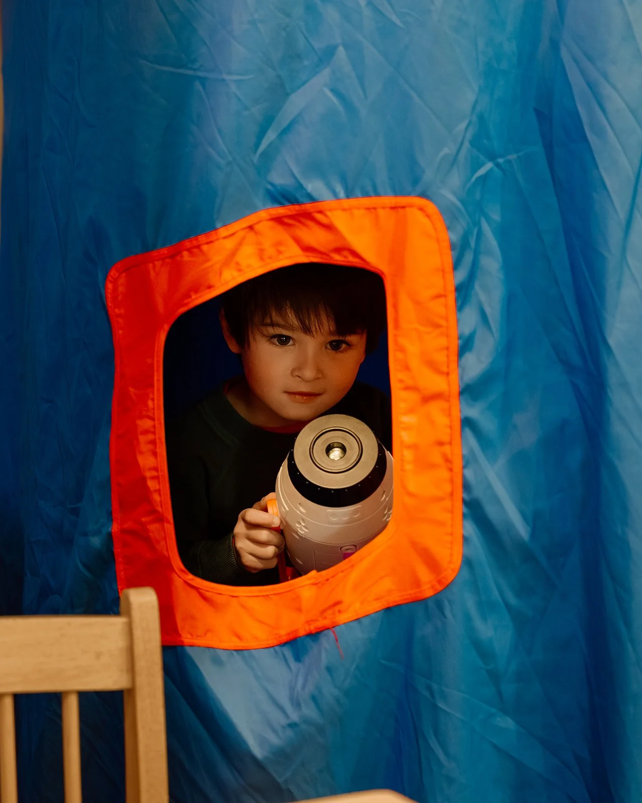 Child looking through a tent window, holding a toy laser gun, with a blue tent and orange trim around the window in the background.